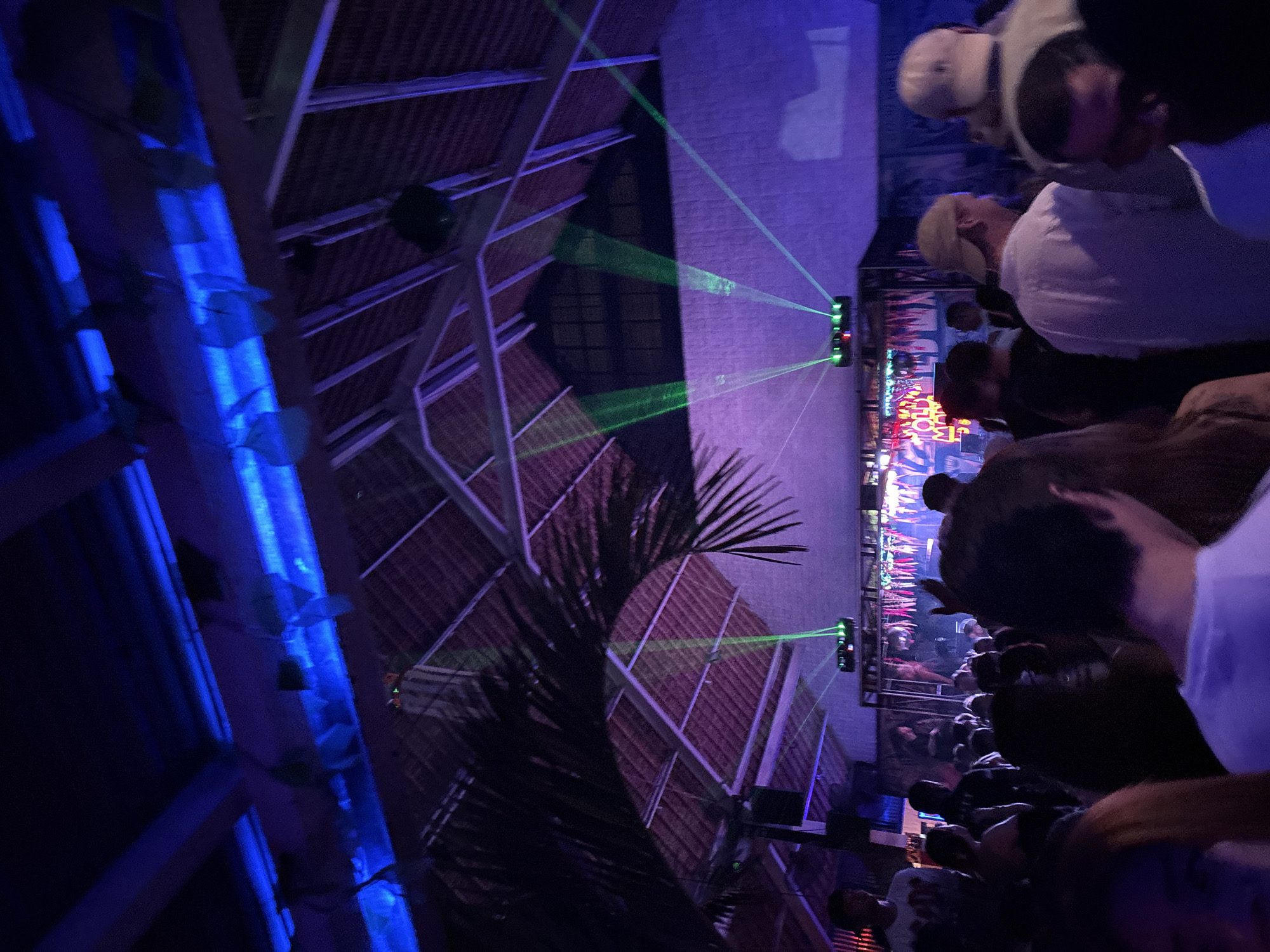 The image depicts a crowded indoor event, likely a party or concert, at night. The venue appears to have a thatched roof structure with exposed wooden beams and is illuminated by a combination of blue and purple stage lighting, with green laser beams projected from a device towards the ceiling and walls. Palm fronds are visible, suggesting a tropical or outdoor-adjacent setting. A diverse group of people, dressed casually, are standing and interacting within the space. In the background, there is a stage area with colorful decorations and what appear to be light fixtures. The overall atmosphere is one of vibrant nightlife. The location is specified as North Kuta, Indonesia.