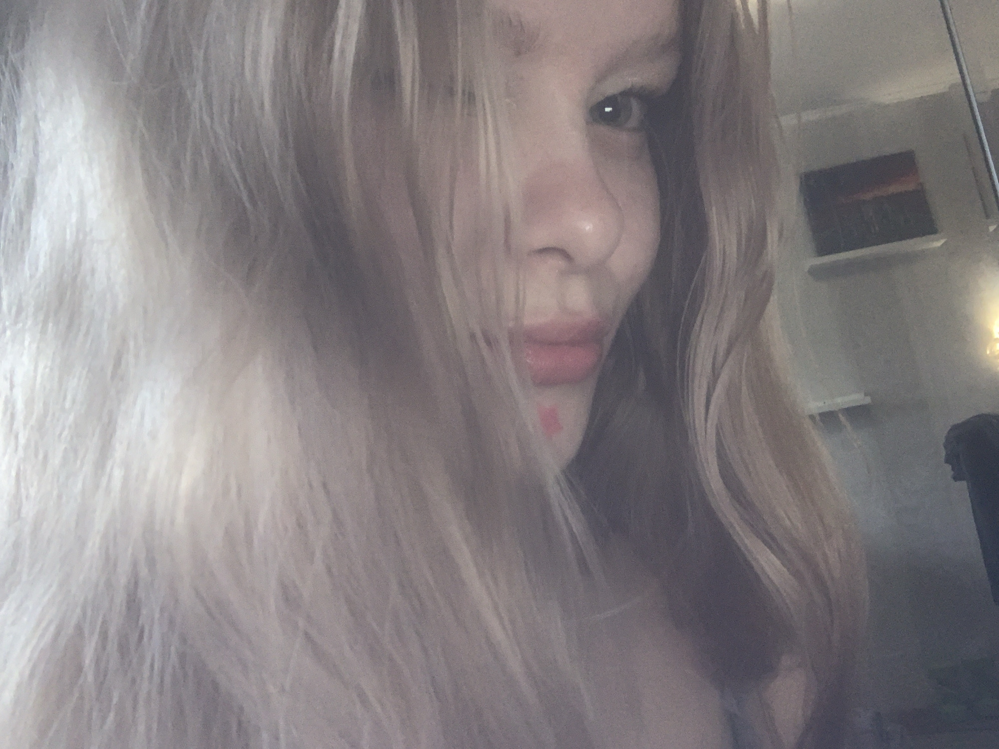 A close-up portrait features a young person, likely female, with fair skin and long, wavy, light blonde hair. The hair cascades down, partially obscuring the left side of the face and the forehead. The person's right eye is clearly visible and looks directly towards the viewer, conveying a neutral or slightly contemplative expression. Visible facial features include the nose and lips, with a small reddish mark or smudge near the right corner of the mouth.
The background suggests an indoor setting, with light-colored walls. On the right side of the frame, behind the person's shoulder, a small rectangular painting or picture depicting a dark landscape or abstract art, possibly a sunset, rests on a narrow white shelf. Further to the right, a darker, indistinct form, possibly furniture or a doorway, is visible, along with a bright light source or reflection. The lighting is soft and diffused, indicating ambient light, likely natural, contributing to a serene atmosphere. No specific activity is depicted beyond posing for the photograph. There is no visible text. The location is an unspecified indoor setting, with no overt cues to indicate the stated context of Russia.
