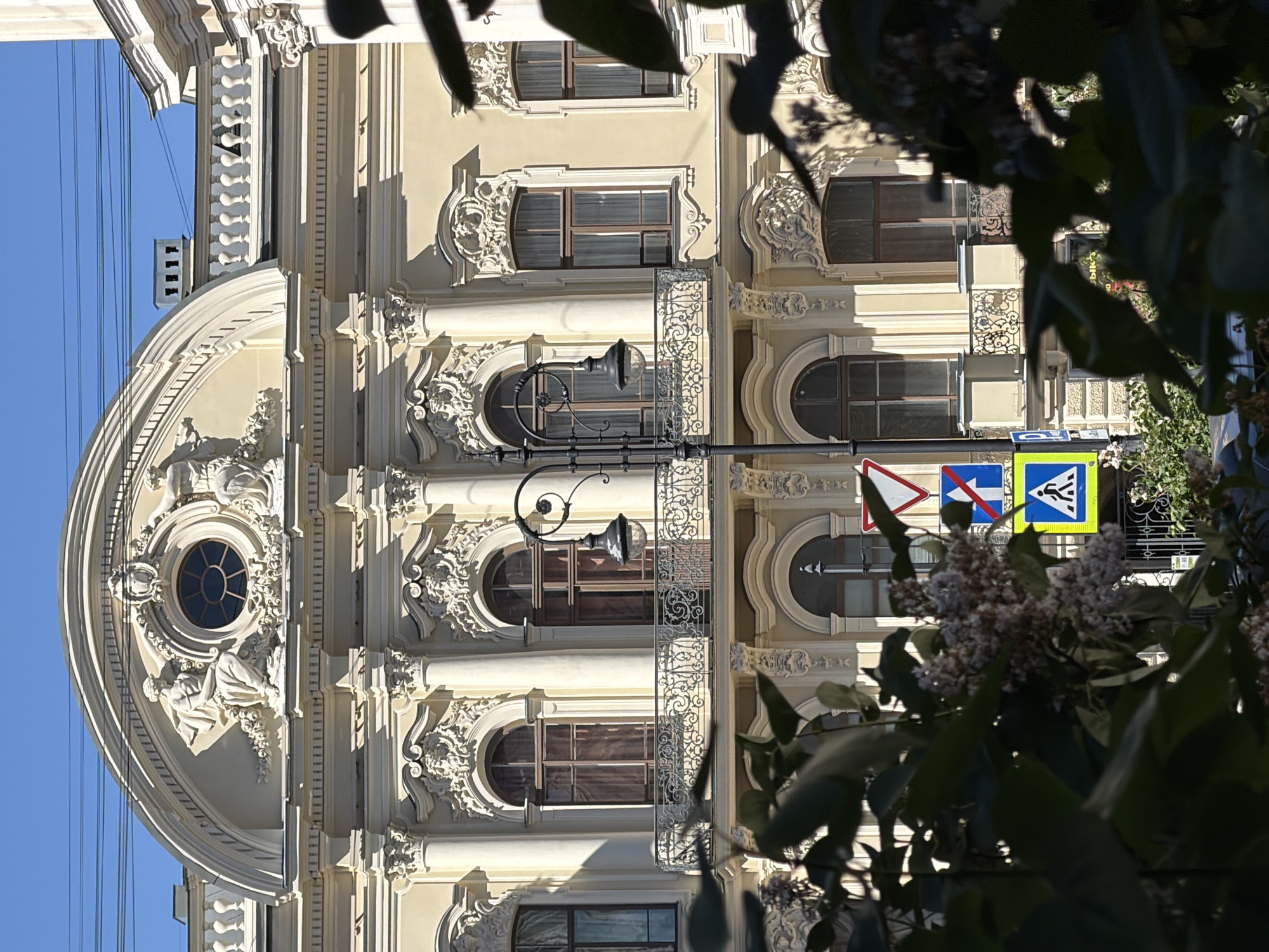 This image captures a segment of an ornate, multi-story building facade in Saint Petersburg, Russia, under a clear blue sky. The building, constructed from light-colored (cream or pale yellow) material, exhibits a classical architectural style with extensive Baroque or Rococo ornamentation.
The uppermost visible section features a large semicircular arch, centrally encompassing a round oculus window. On either side of the oculus, two relief figures, depicted in classical drapery, are seated. The figure on the left is fully discernible, while the one on the right is partially obscured. Above the oculus, an ornate cartouche is present.
The main facade displays multiple tall, arched windows framed in dark brown, with light-colored curtains visible through the panes. These windows are interspersed with engaged columns featuring elaborate Corinthian-style capitals. Rich stucco decorations, including mascarons and floral patterns, are meticulously applied above and between the windows. A decorative wrought-iron balcony with intricate scrollwork extends across a portion of this level, beneath two central windows.
The lower section of the building, partially obscured by foreground elements, continues the architectural theme with additional arched windows and detailed stucco work. A dark metal gate is faintly visible on the right at ground level.
In the mid-ground, a black, ornate street lamp post with two spherical glass globes stands, partially obstructing the view of the building. One of the lamp globes appears to have a white, frosty, or dusty coating. Several thin, dark electrical wires traverse the upper portion of the clear blue sky.
The lower foreground is dominated by dense, dark green foliage interspersed with clusters of small, light-colored blossoms, which obscure the building's base and ground-level elements. Through this foliage, several Russian traffic signs are partially visible: a red-bordered white triangular "Give Way" sign, a blue rectangular "No Straight Ahead" sign, a blue square "Pedestrian Crossing" sign, and a partially visible blue "P" for parking sign. No individuals are present in the depicted scene.