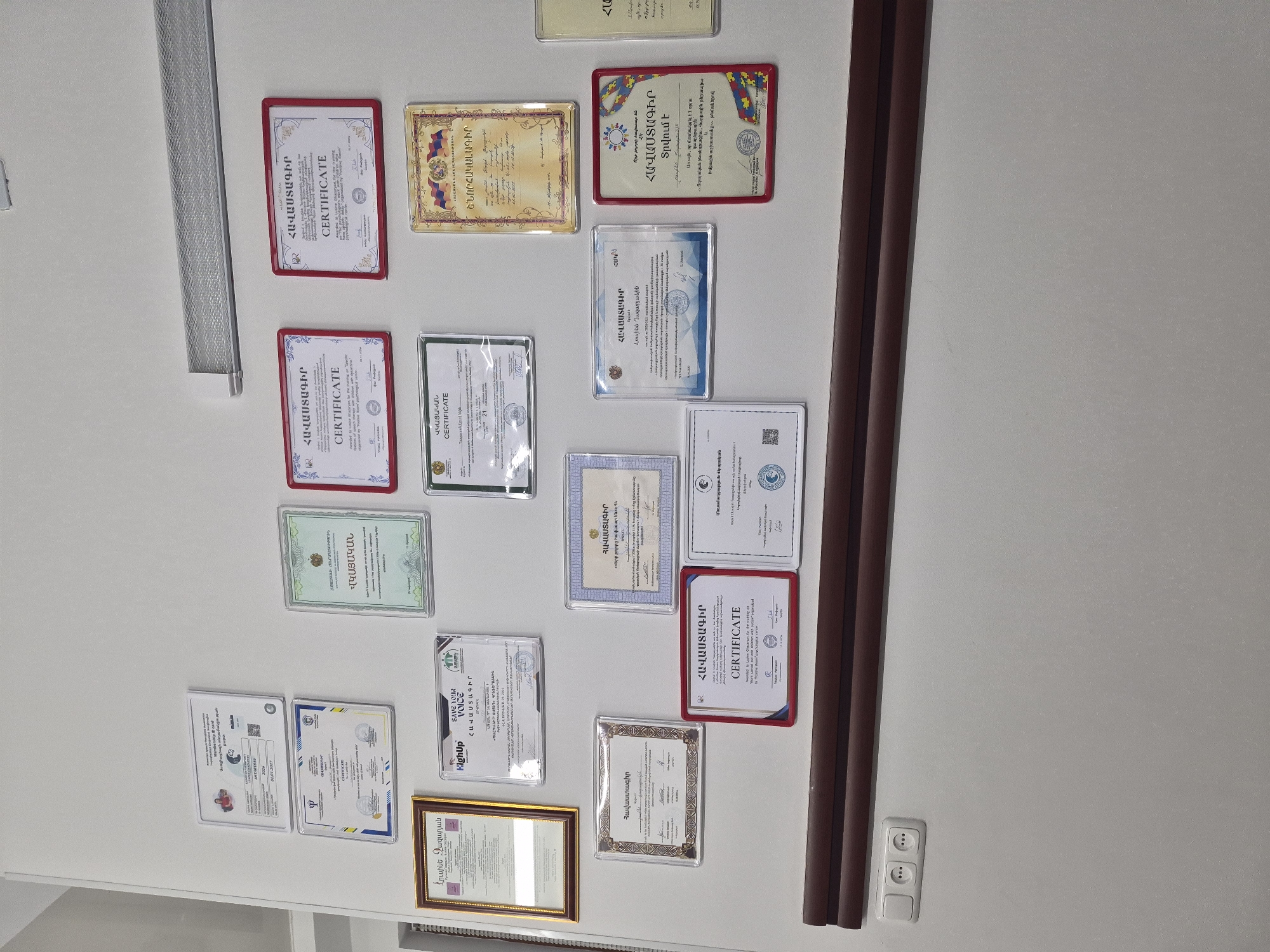 The image shows a wall adorned with numerous framed certificates, predominantly in Armenian. The certificates appear to be official documents, possibly academic or professional achievements, and are displayed in various sizes and frames, some with red borders and others with gold or simple clear plastic sleeves. The wall is painted a light, neutral color, and a dark brown trim runs horizontally across the lower portion of the image. To the left of the trim, a light switch and two electrical outlets are visible. The certificates are arranged in a somewhat organized, yet dense, manner.
Based on the visible Armenian script on the certificates and the context of such displays often found in professional settings, this image is likely from a location in Yerevan, Armenia. Without specific identifiable landmarks or further contextual clues within the image, pinpointing an exact street or building is not possible, but it is consistent with an office, clinic, or educational institution in the city. No people are visible in the image.