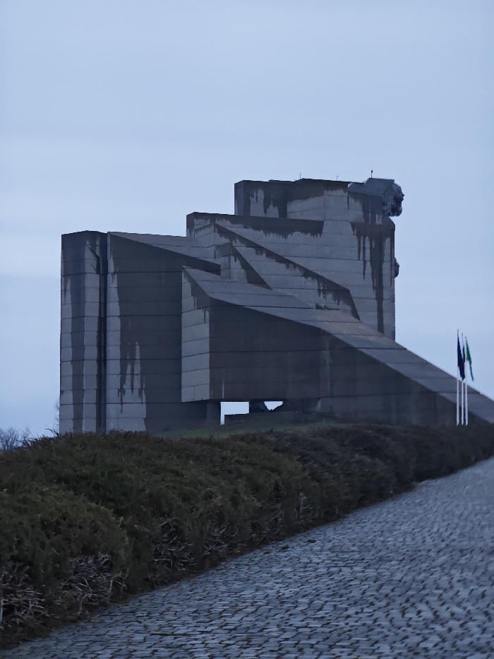 The image displays the Monument to 1300 Years of Bulgaria, located on the Shumen Plateau overlooking Shumen, Bulgaria. The monument is a large, brutalist concrete structure composed of angular, geometric blocks arranged in a stepped and zig-zagging configuration. Dark vertical staining is present on the grey concrete surfaces. A sculptural element, appearing as a stylized head or face, is partially visible on the upper right section of the monument. In the lower right foreground, a cobblestone pathway ascends, bordered on its left by a dense row of dark green shrubs. Three flagpoles are positioned to the right of the monument, two of which bear flags: one dark blue and one green. The sky is uniformly light grey and overcast. No individuals, animals, or active subjects are present.