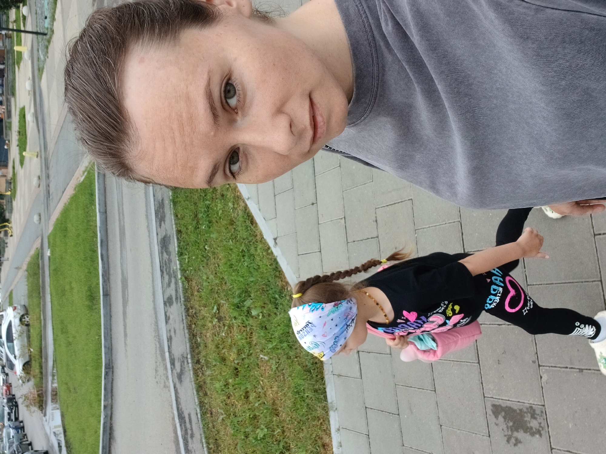 A woman and a young girl are walking on a sidewalk in Yekaterinburg, Russia. The woman is in the foreground, taking a selfie. She has brown hair and is wearing a gray t-shirt. The girl is walking next to her, wearing a black t-shirt with colorful graphics, black leggings with a pink heart, a colorful bandana, and carrying a pink bag. She has a braid and a beaded necklace. In the background, there are cars parked along a street and a patch of grass. The weather appears to be overcast.
