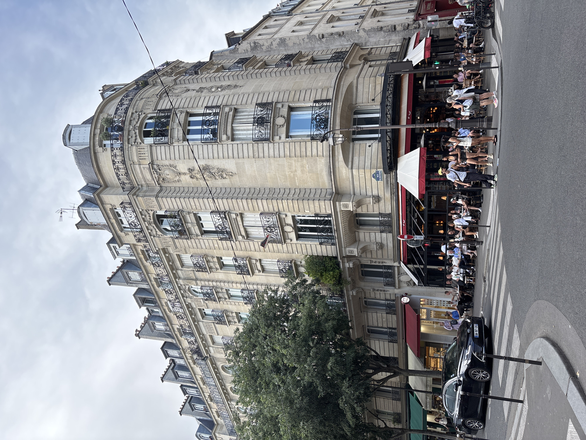 The image depicts a corner building in Paris, France, with a street-level cafe and shops. The building is made of light-colored stone with ornate architectural details, including balconies with wrought-iron railings and mansard roofs. The cafe has red and white awnings and outdoor seating where several people are dining and socializing. A black sedan is driving on the street, and pedestrians are crossing at a crosswalk. A large tree stands on the sidewalk, providing shade. The sky is overcast.