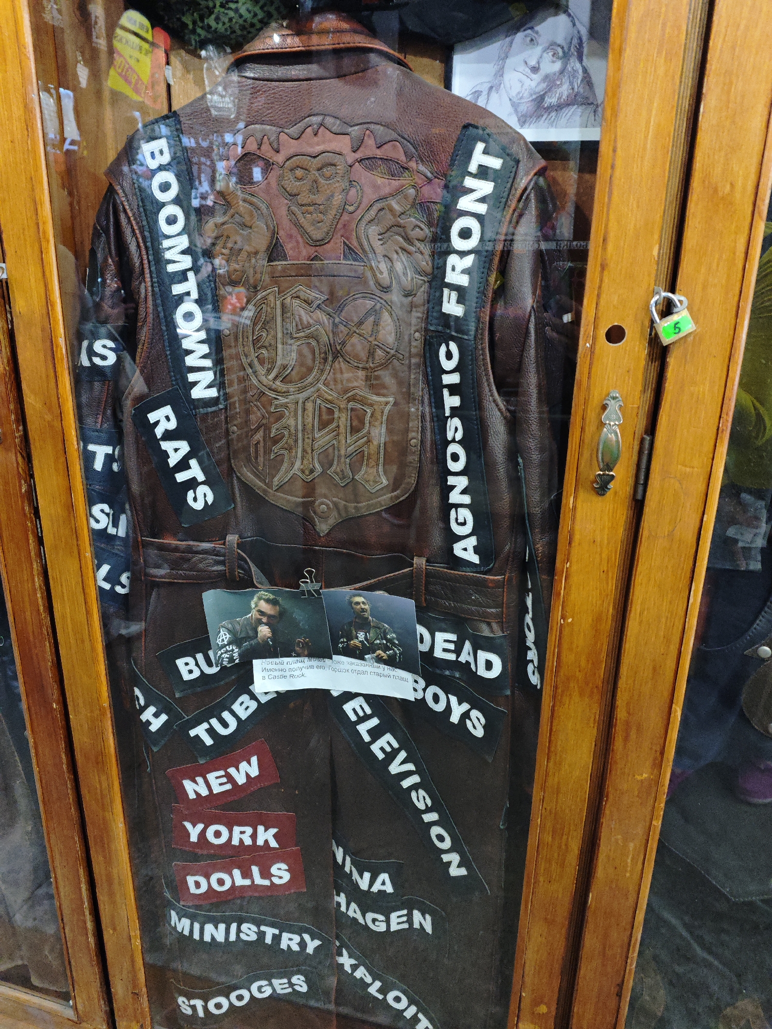The image shows a close-up view of a brown leather jacket adorned with numerous patches, displayed in a glass case with wooden framing. The jacket features various band names and symbols stitched onto it. Prominent patches include "BOOMTOWN," "RATS," "AGNOSTIC FRONT," "DEAD BOYS," "TELEVISION," "NEW YORK DOLLS," "MINISTRY," and "STOOGES." There is also a central emblem with intricate detailing and what appears to be lettering. Two small photographs are attached to the jacket, one featuring a man speaking into a microphone. A small padlock is visible on the right side of the wooden frame, with a green tag bearing the number "5". The background is partially visible, showing a sketch of a face and some other items within the display case. This jacket is part of an exhibit at the Museum of Russian Rock Music (Музей русского рок-н-ролла) in Saint Petersburg, Russia.