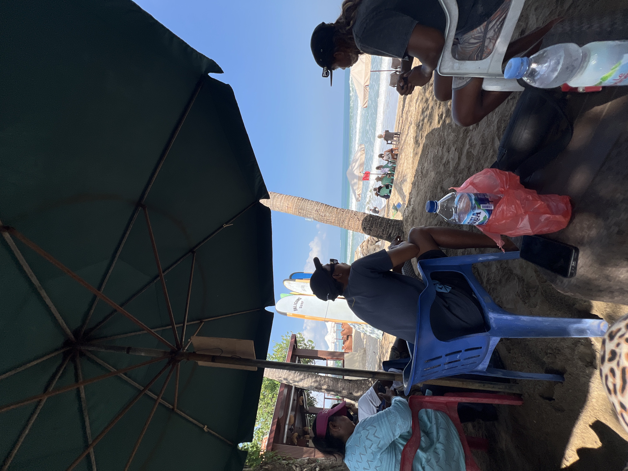 The image depicts a beach scene in Kuta, Indonesia, under a large green umbrella providing shade. In the foreground, three individuals are seated on chairs. To the left, a person wearing a blue top and a pink visor is seated on a red chair. In the center, a person wearing a dark t-shirt and a black baseball cap is seated on a blue plastic chair, facing away from the camera. To the right, another person in a dark t-shirt and a black baseball cap is seated on a white chair, also facing away from the camera.
On the sand, in front of the central figure, a black smartphone and a red plastic bag containing a water bottle are visible. A second water bottle is positioned in the lower right corner of the frame.
In the midground, the sandy beach extends towards the ocean. Several surfboards are propped up near a beachside structure. Further along the beach, more people are seated under umbrellas. The ocean with gentle waves and a clear blue sky are in the background. The overall environment suggests a relaxed beach day in Kuta.