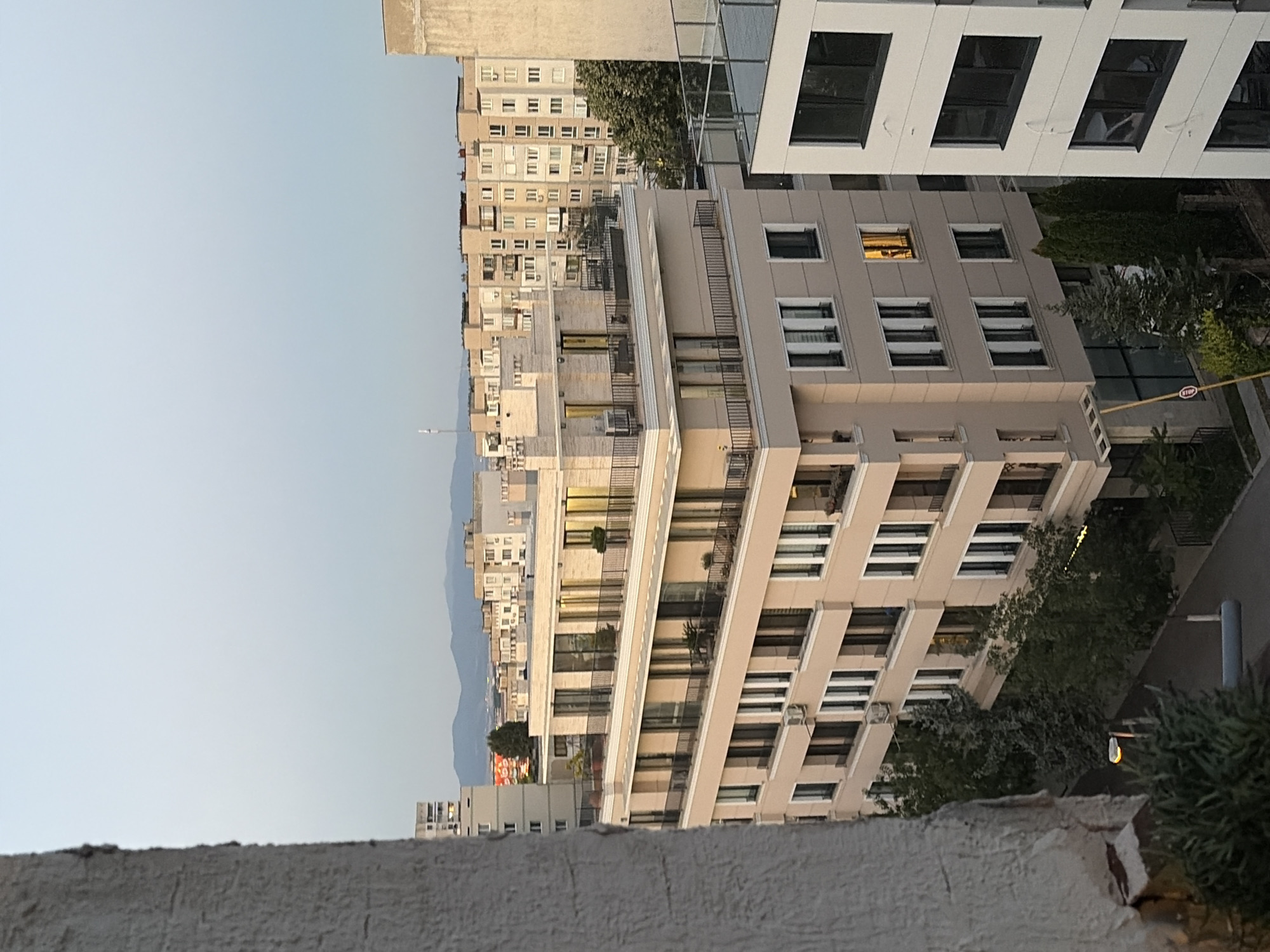 An exterior, eye-level, wide shot shows a row of modern, beige apartment buildings in a city on a clear day. In the foreground, to the left, is the rough, textured, grey outer wall of a building, partially obscuring the view of the other buildings. The main focus is on a multi-story beige building with many windows and balconies. The building has a flat roof with black railings and some potted plants. The windows on the upper floors are illuminated with warm, yellow light, and one window on the fifth floor shows a silhouette of a person standing in front of the light. There are several other apartment buildings of varying heights and styles in the background, some with visible balconies and windows. In the distance, hazy mountains are visible against a pale blue sky. Green trees are planted between the buildings and along the street, some of which are tall and slender, while others are shorter and bushy. A paved street runs in front of the buildings, with a street light and some signage visible. The overall scene depicts an urban residential area with a sense of twilight or late afternoon light.