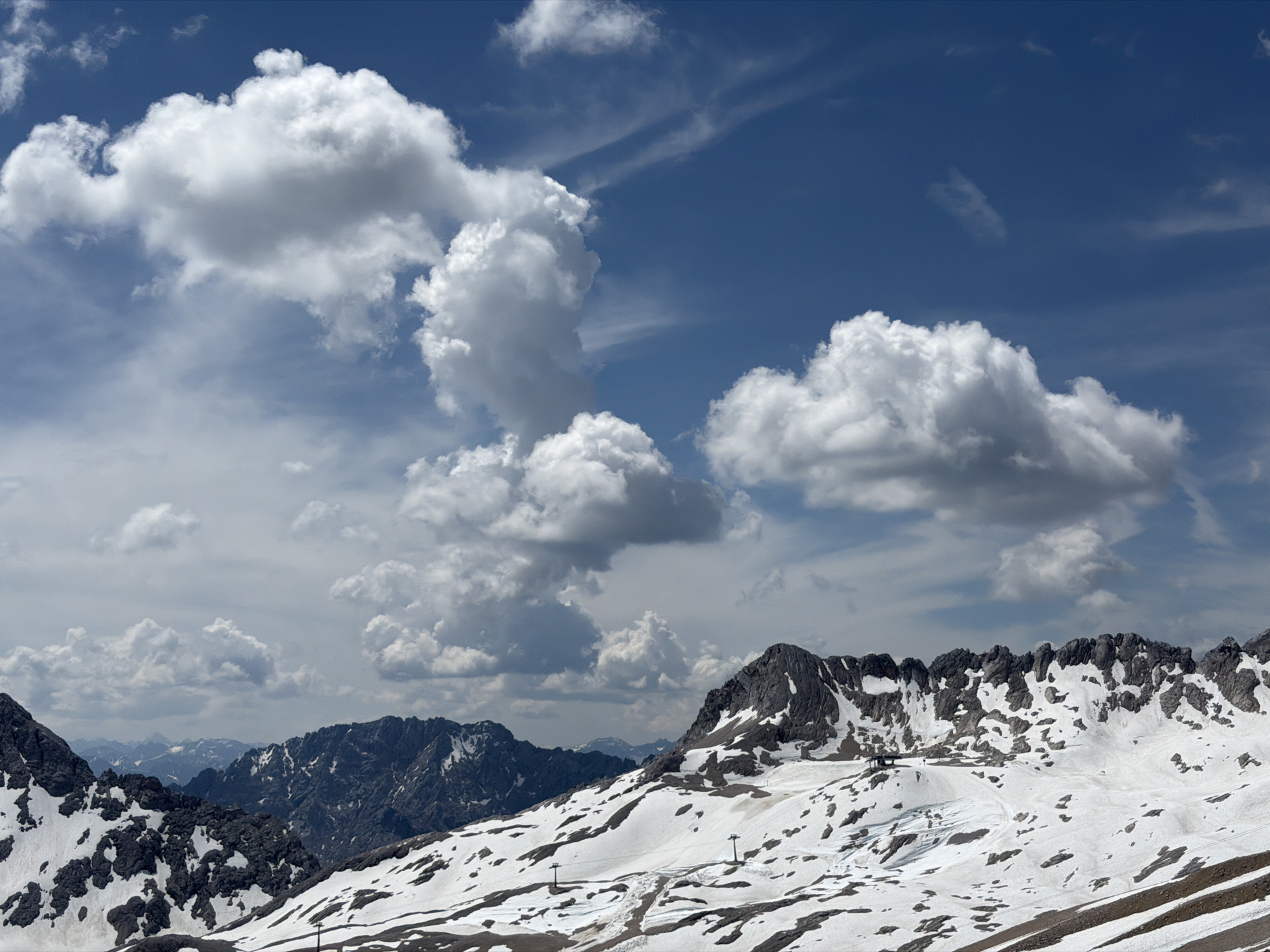 The image depicts a panoramic view of a snow-capped mountain range under a partly cloudy blue sky. The foreground is dominated by a rugged mountain slope with patches of snow and exposed dark rock. Several ski lift pylons are visible on the slope, suggesting a ski resort or a popular hiking area. In the mid-ground, more jagged mountain peaks rise, also partially covered in snow. The background features a vast expanse of distant mountains fading into a hazy blue. The sky is a vibrant blue, punctuated by fluffy white cumulus clouds and thinner, wispy cirrus clouds. The overall scene conveys a sense of altitude and a cool, crisp mountain atmosphere. This landscape is characteristic of the Alps in the vicinity of Garmisch-Partenkirchen, Germany, likely captured from a high vantage point such as the Zugspitze or a nearby summit, offering views of the surrounding alpine terrain. No people are visible in the image.