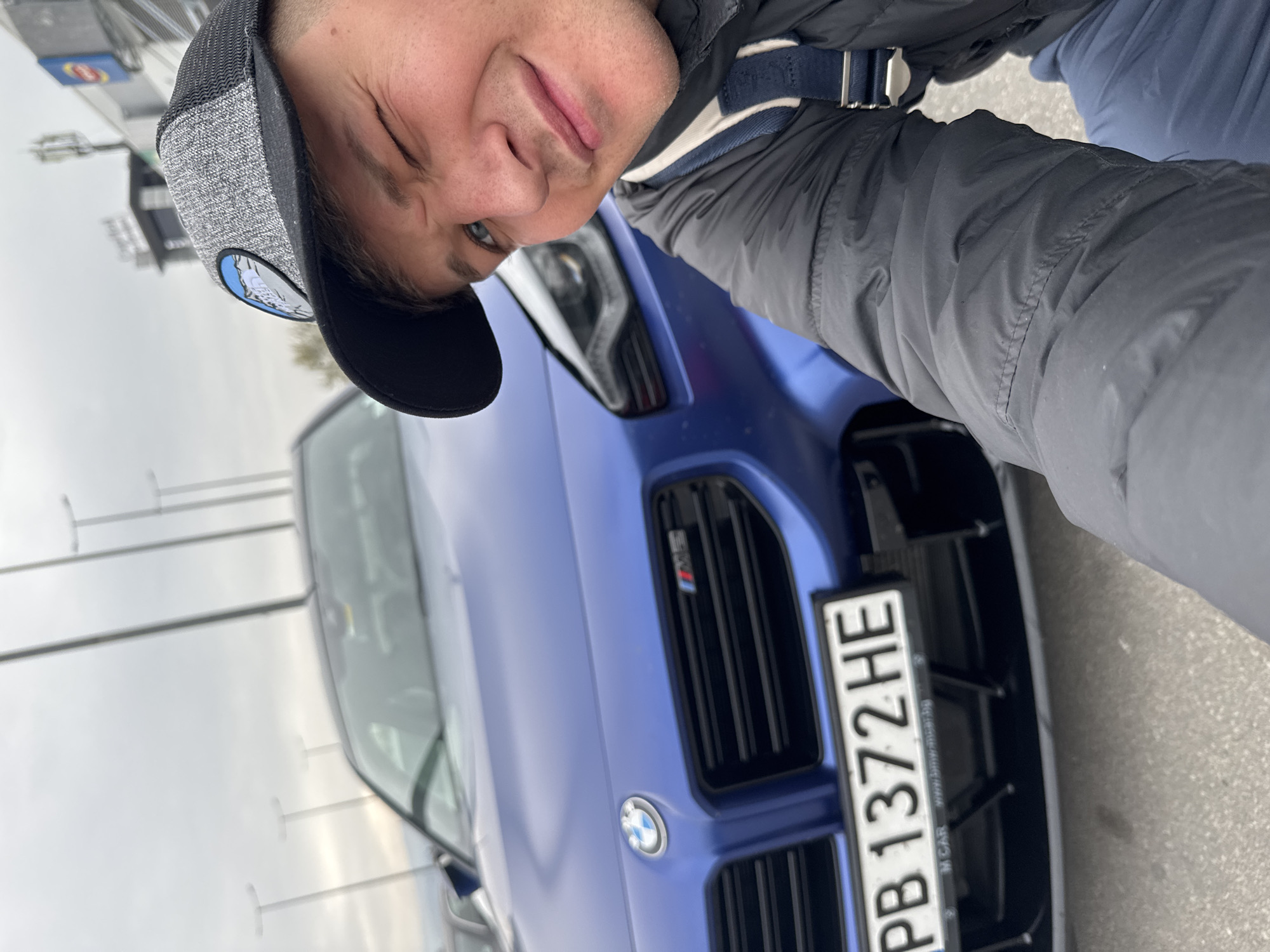 A man in a grey cap and black jacket is standing next to a blue BMW car. The car's license plate reads "PB 1372 HE." The sky is overcast. The location is possibly in Plovdiv, Bulgaria, based on the license plate.