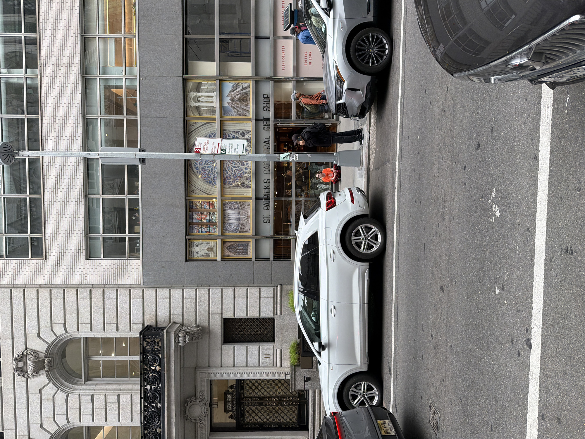 The image depicts a street scene in New York City, specifically in front of St. Patrick's Cathedral's Central Gift Shop. Several vehicles are parked along the side of the road, including a white SUV and a gray sedan. The architecture of the buildings is visible, showcasing a mix of brick and stone facades with large windows and ornate details. Pedestrians are present, including individuals standing and walking in front of the gift shop. A traffic sign indicating restrictions for commercial vehicles is visible. The street itself appears to be paved asphalt with lane markings.