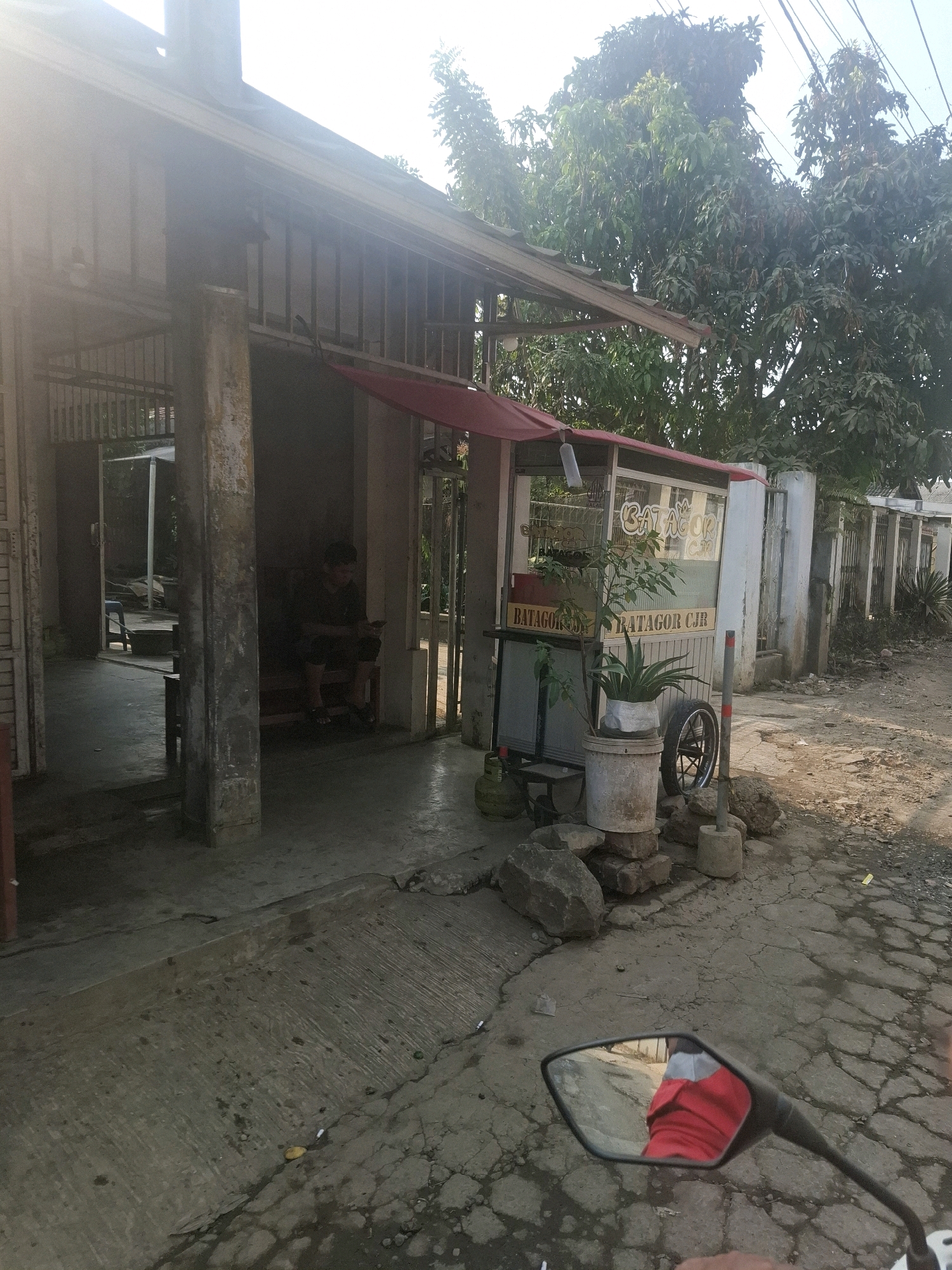 The image shows a street scene in Ciheulang Tonggoh, Indonesia. In the foreground, a motorcycle mirror reflects a person wearing a red shirt. The road is cracked and appears to be paved with asphalt. To the right, there is a food stall with the sign "BATAGOR CJR". Various items are placed around the stall, including rocks, a spare tire, and two potted plants. Behind the stall, a fence runs along the property line, and trees are visible in the background. To the left of the stall, an open doorway leads into a dimly lit interior space where a person is sitting on a bench.