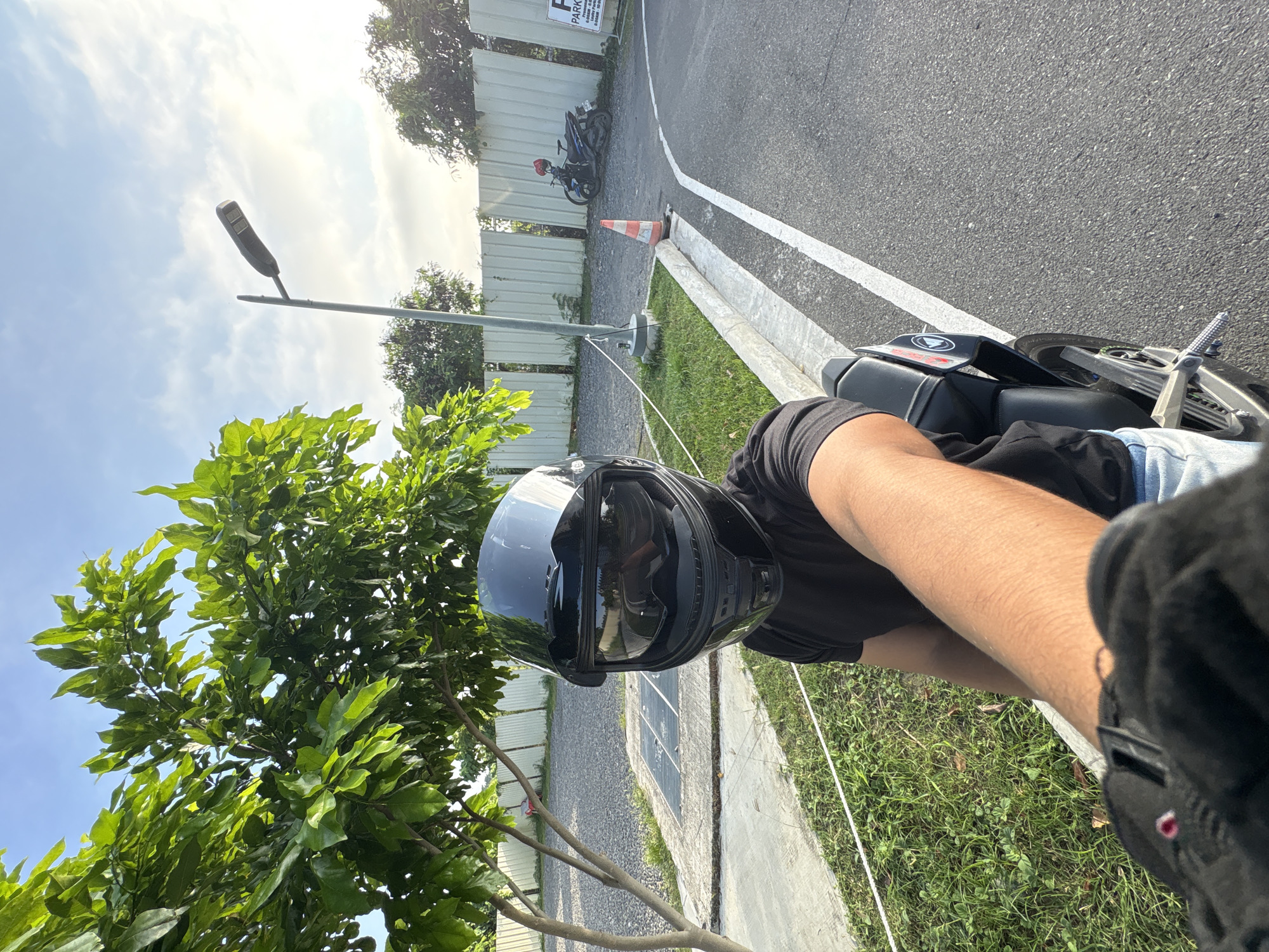 A first-person perspective selfie shows a person wearing a black helmet and black t-shirt, sitting on a motorcycle. The person's left arm and hand, covered by a black glove, are visible in the foreground. The background includes a tree with green leaves, a gravel area, a concrete path, grass, a white metal fence, a lamppost, a traffic cone, and a blue motorcycle with a red helmet parked near a sign indicating parking. The scene appears to be outdoors, possibly in a parking lot or driving range, with a bright sky. The presence of a parking sign and the general layout suggest an area designed for vehicles. Given the sunny weather and foliage, it is likely daytime. The location in Davao City, Philippines, can be inferred from similar scenes found in local establishments and training grounds.