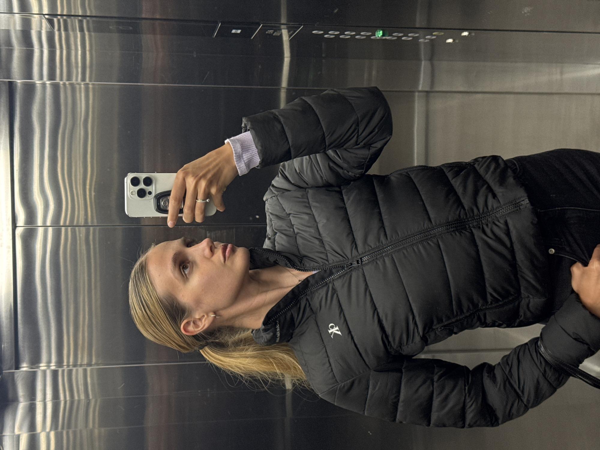 A woman stands inside an elevator, likely taking a selfie with a white smartphone. She is wearing a black puffer jacket with a "CK" logo visible on the collar, and a lavender-colored shirt under the jacket. Her blonde hair is pulled back into a low ponytail. The elevator interior appears to be made of brushed metal, with the control panel and floor indicator lights visible in the background. The location in the city of Unknown, Unknown cannot be determined from the image.