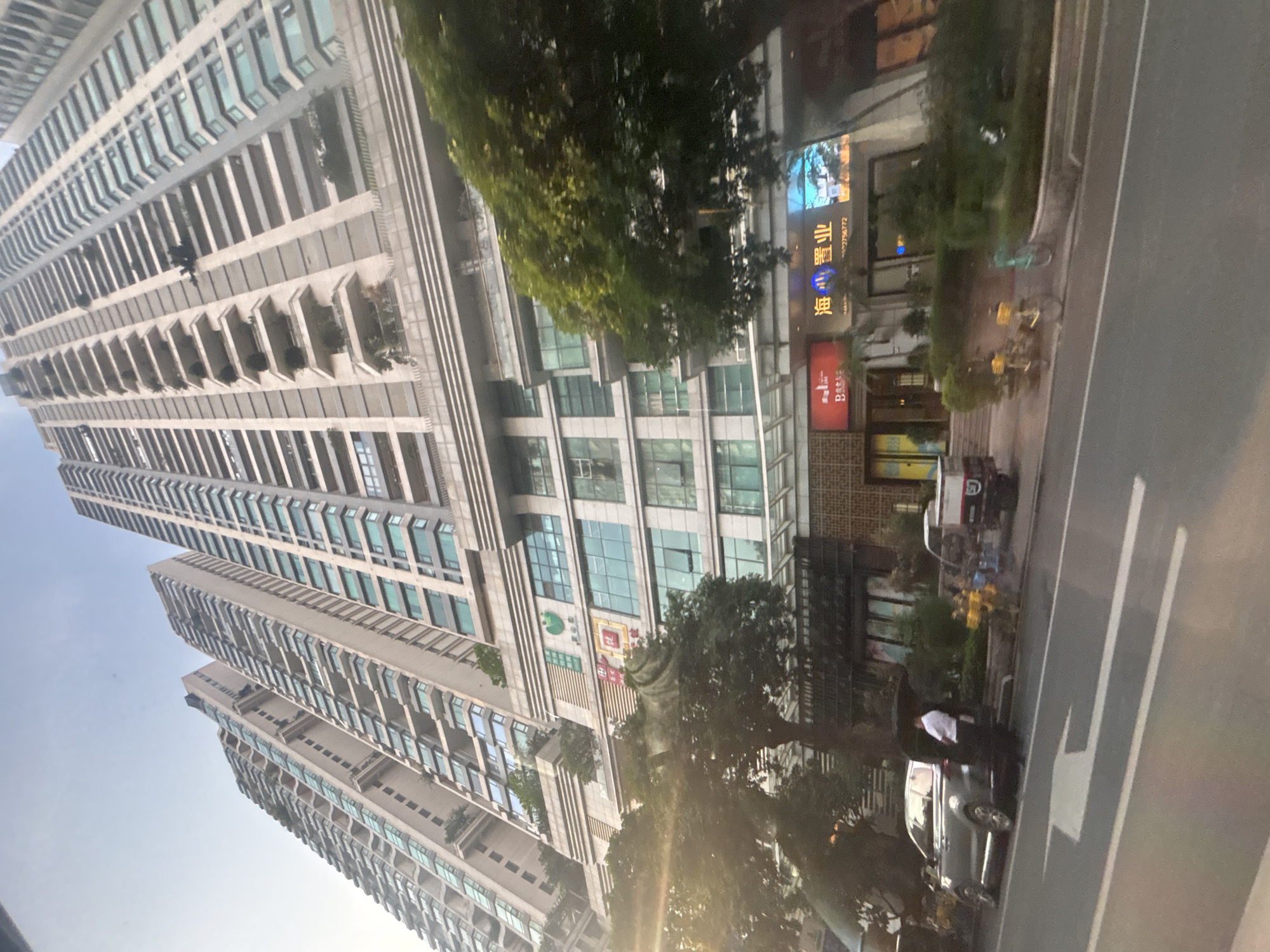 The image captures a wide shot of a modern cityscape, with towering residential buildings dominating the background. The buildings are made of light-colored concrete and feature numerous balconies with glass railings. In the foreground, a street is visible, with a silver SUV parked on the side. A person in a white shirt and dark pants stands next to the open trunk of the SUV. Adjacent to the building are several shops, one with a red awning and Chinese characters, and another with a prominent illuminated sign displaying Chinese characters that read "海 碧 业". There are also several parked bicycles and a small three-wheeled vehicle with a white box on the back. The environment suggests a bustling urban setting, likely in Haizhu District, China, given the signage. The lighting indicates it could be late afternoon or early evening, with the sun casting a slight glare on the left side of the image.