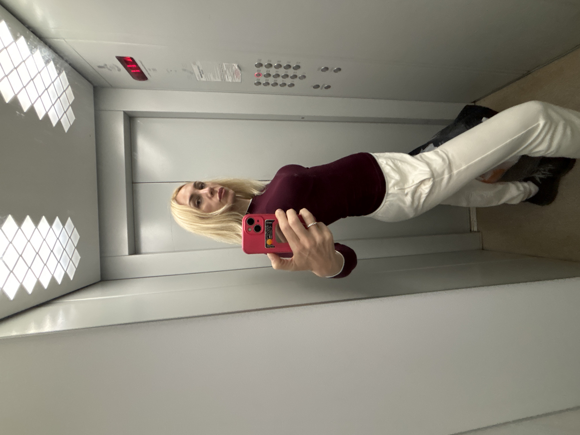 A woman with blonde hair is taking a mirror selfie in an elevator. She is wearing a maroon long-sleeved shirt and loose white pants. She is holding a pink smartphone with a credit card attached to the case in her right hand. The elevator has grey walls and a tiled ceiling light. There is an elevator control panel with buttons and a digital floor indicator displaying "114". The floor of the elevator is a neutral beige color. The image does not provide enough information to determine the specific location within the city of Imeni Morozova, Russia.