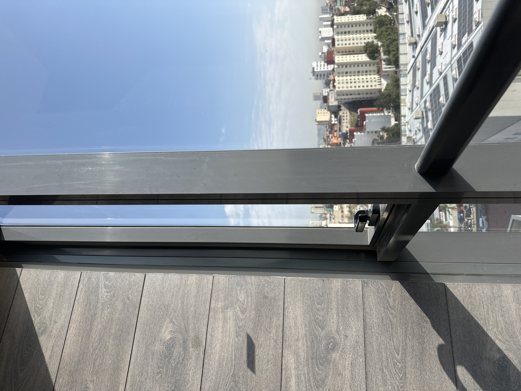 The photograph shows an elevated perspective of a cityscape from a high-rise building in Huixquilucan de Degollado, Mexico. The scene is viewed through a partially opened window, with a grey metal frame and locking mechanism visible in the foreground. Outside the window, the buildings of the city are visible, including white high-rise buildings. The sky is pale blue. The floor inside is wooden. The image provides an upward angle view to the exterior environment.