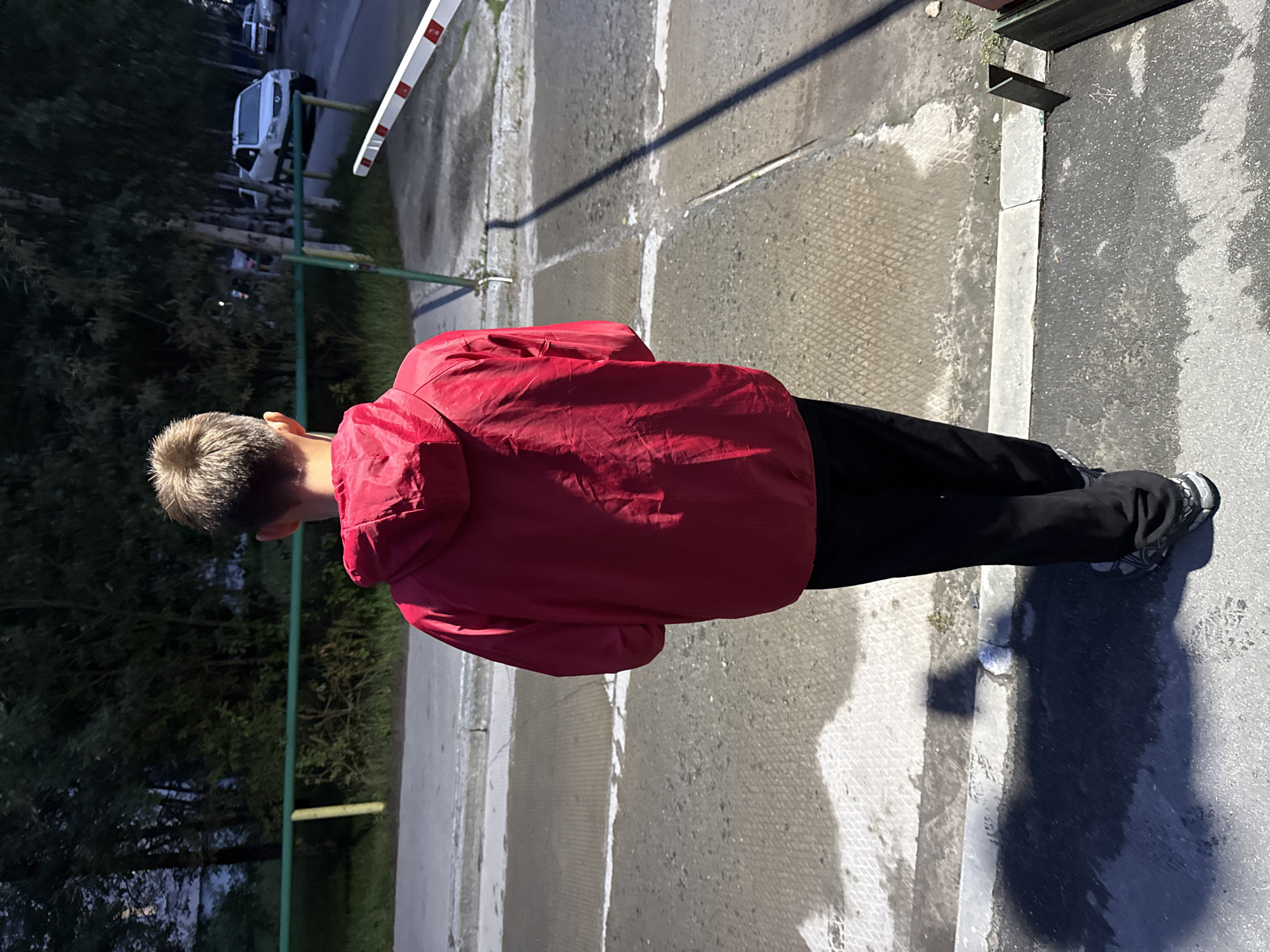 A young boy, seen from the back, stands on a paved surface next to a barrier. He is wearing a bright red hooded jacket and black pants. The background consists of trees and several parked cars, including a white SUV. A white and red barrier arm extends into the frame from the right. The ground is a mixture of asphalt and concrete, with some puddles of water creating reflections. A shadow stretches across the ground from the boy and a vertical pole. The lighting suggests it is either early morning or late evening.