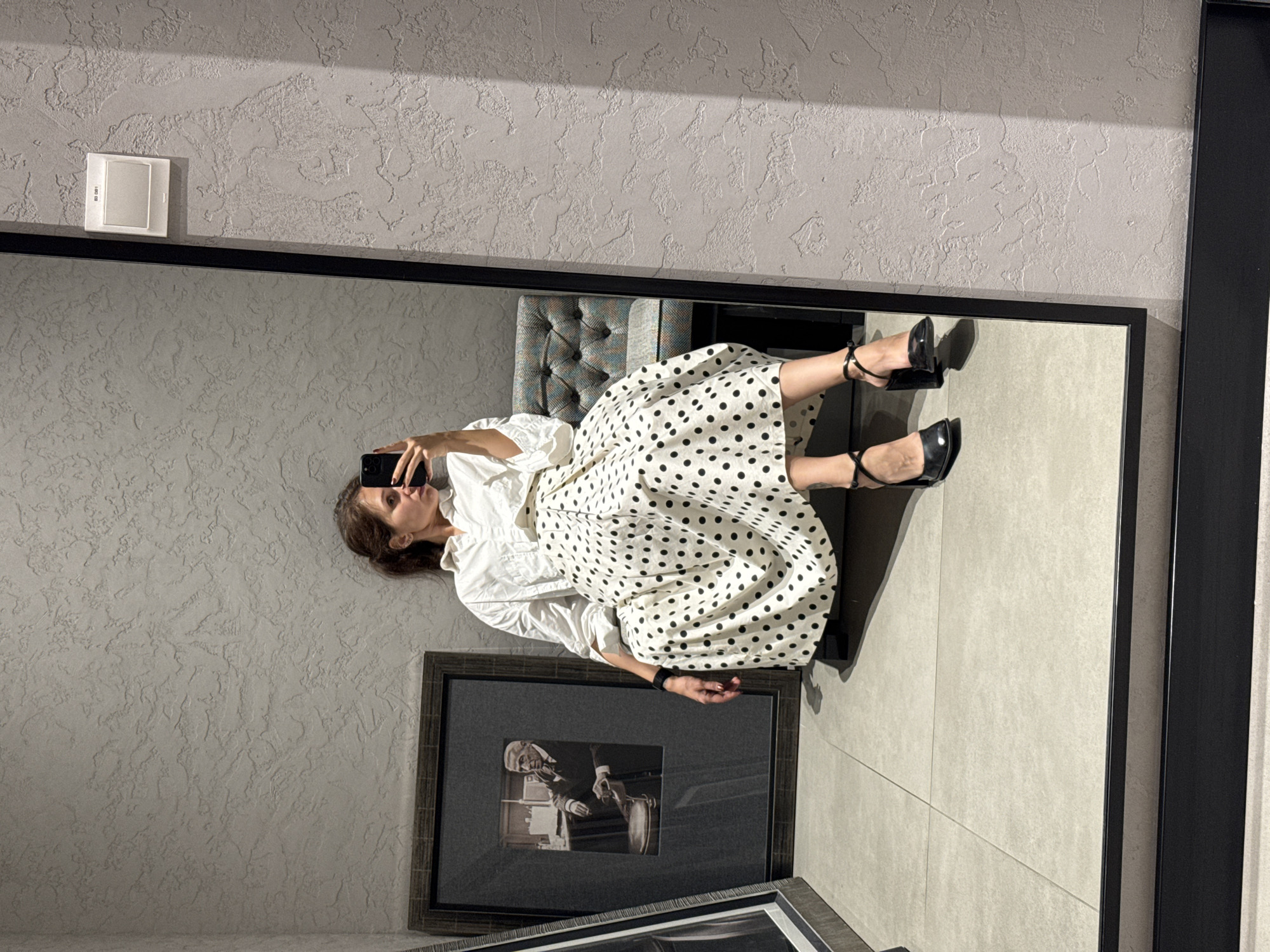 The image presents a mirror selfie of a woman within a modern interior setting. The woman, with brown hair, is seen from her upper body to her legs in the reflection. She is dressed in a crisp white long-sleeved blouse and a cream-colored skirt patterned with black polka dots. Her feet are adorned with black high-heeled shoes featuring ankle straps, and she wears a dark-colored smartwatch or bracelet on her left wrist. She holds a black smartphone to take the selfie, with her gaze directed towards the phone screen.
She appears to be seated or leaning back against a partial view of a tufted armchair or sofa, upholstered in a light blue and grey patterned fabric. Below her reflection, resting on the floor or a low surface, are two framed pictures, one of which prominently displays a black and white image of an older man in a suit.
The actual room features a light grey, heavily textured wall with a rough plaster finish. A white square wall switch or outlet, marked with "ED 081," is visible on this wall in the upper left. The floor is composed of large, light-colored square tiles. A stark black frame encloses the large mirror. Strong shadows are cast on the textured wall, indicating a significant light source, likely from a window, positioned to the upper right of the scene (when viewed upright). The overall ambiance is clean and contemporary.