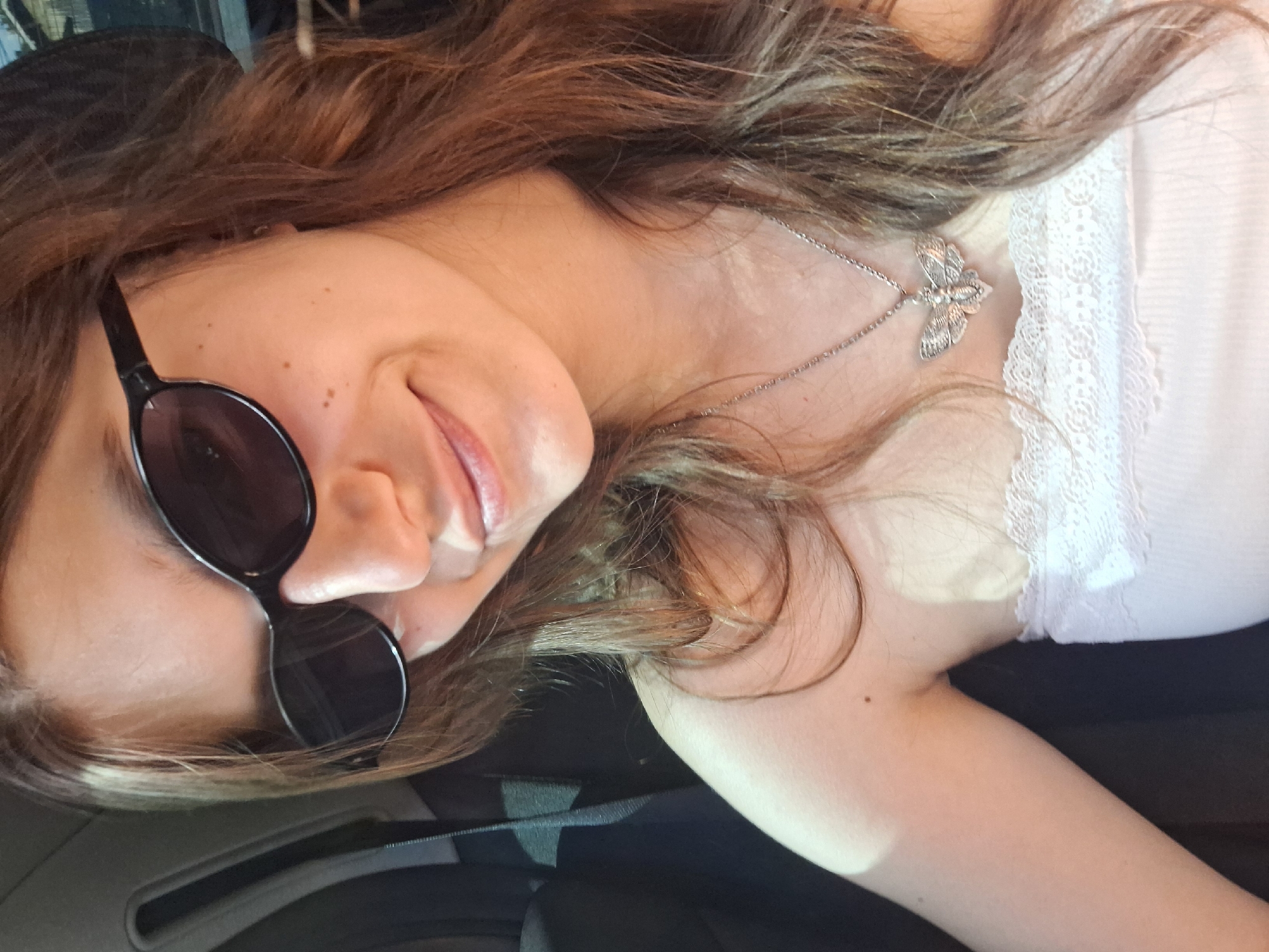 This media file is a close-up, horizontally oriented selfie of a young woman. She is wearing large, dark sunglasses and has her mouth slightly open, revealing her tongue. Her long, brown hair is styled loosely and frames her face. A delicate silver necklace with a dragonfly pendant rests on her décolletage. She is wearing a white top with lace trim. The background appears to be indoors, possibly a car interior, with dark upholstery visible. The lighting suggests it could be daytime, with natural light illuminating her face. There are no visible text elements in the image. The overall mood conveyed is casual and relaxed. The provided location context, Koprivshtitsa, Bulgaria, is not directly evident from visual cues within the image itself but is stated as external information.