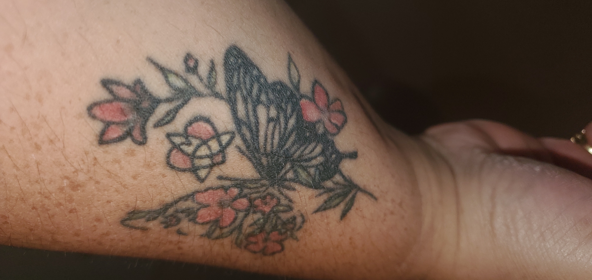 A close-up, low-angle shot shows a tattoo of a butterfly with pink flowers on a person's forearm. The tattoo is rendered in black ink for the butterfly and its floral companions, with accents of pink for the petals of the flowers. Some of the flowers appear to be in bloom, while others are buds. The butterfly's wings are detailed with intricate patterns. The skin of the forearm displays numerous small, brown freckles. The background is dark and out of focus. The image was taken in Sasbach, Germany.