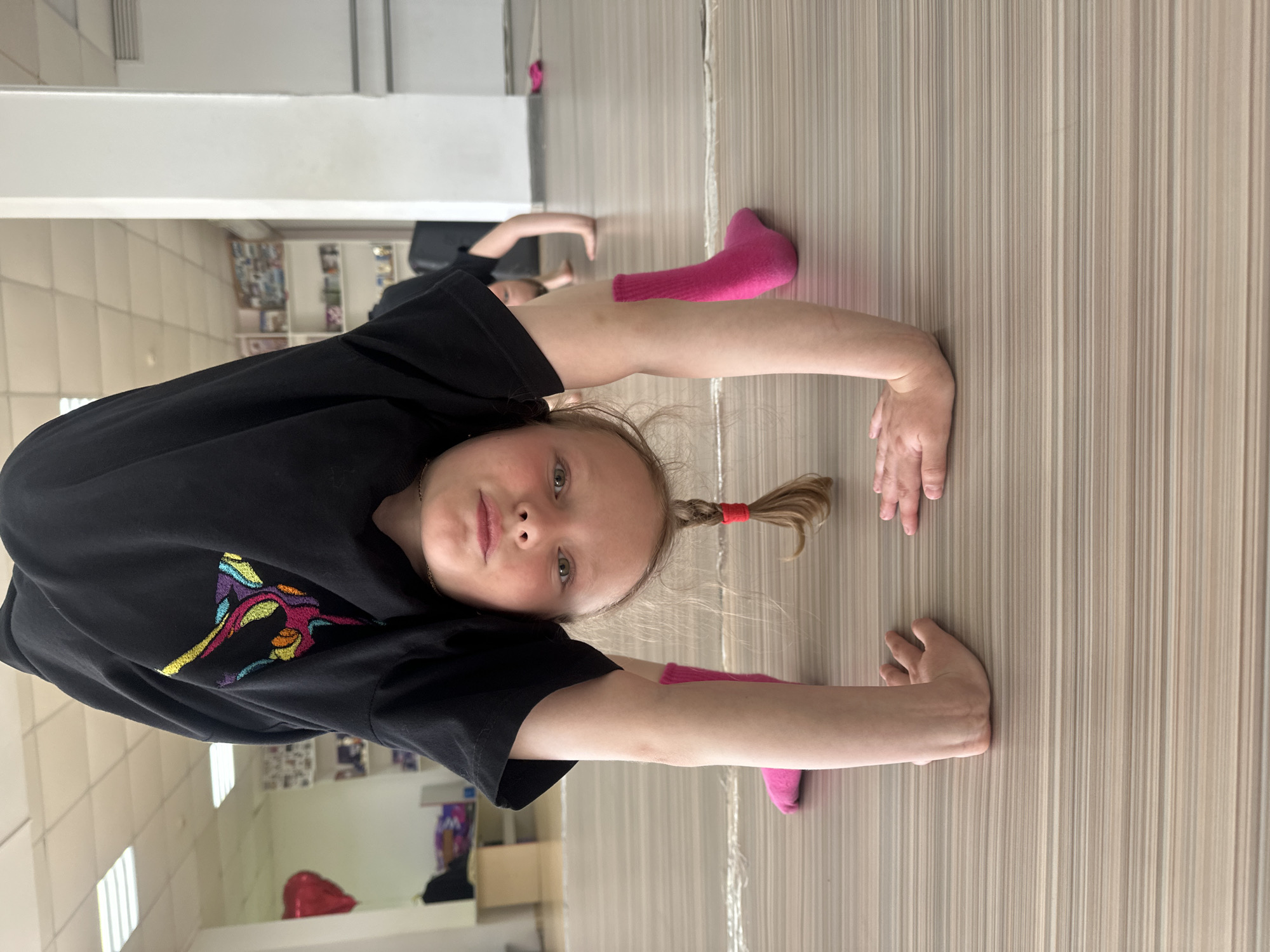 A young girl in a black t-shirt with colorful embroidery is performing a gymnastic stretch on a wooden-look floor. She is wearing pink socks, and her hair is in a ponytail tied with a red band. Behind her, another person is stretching as well. The location appears to be a dance or gymnastics studio, with a mirror on the left and a glimpse of a cityscape out the window. The city is likely Votkinsk, Russia.