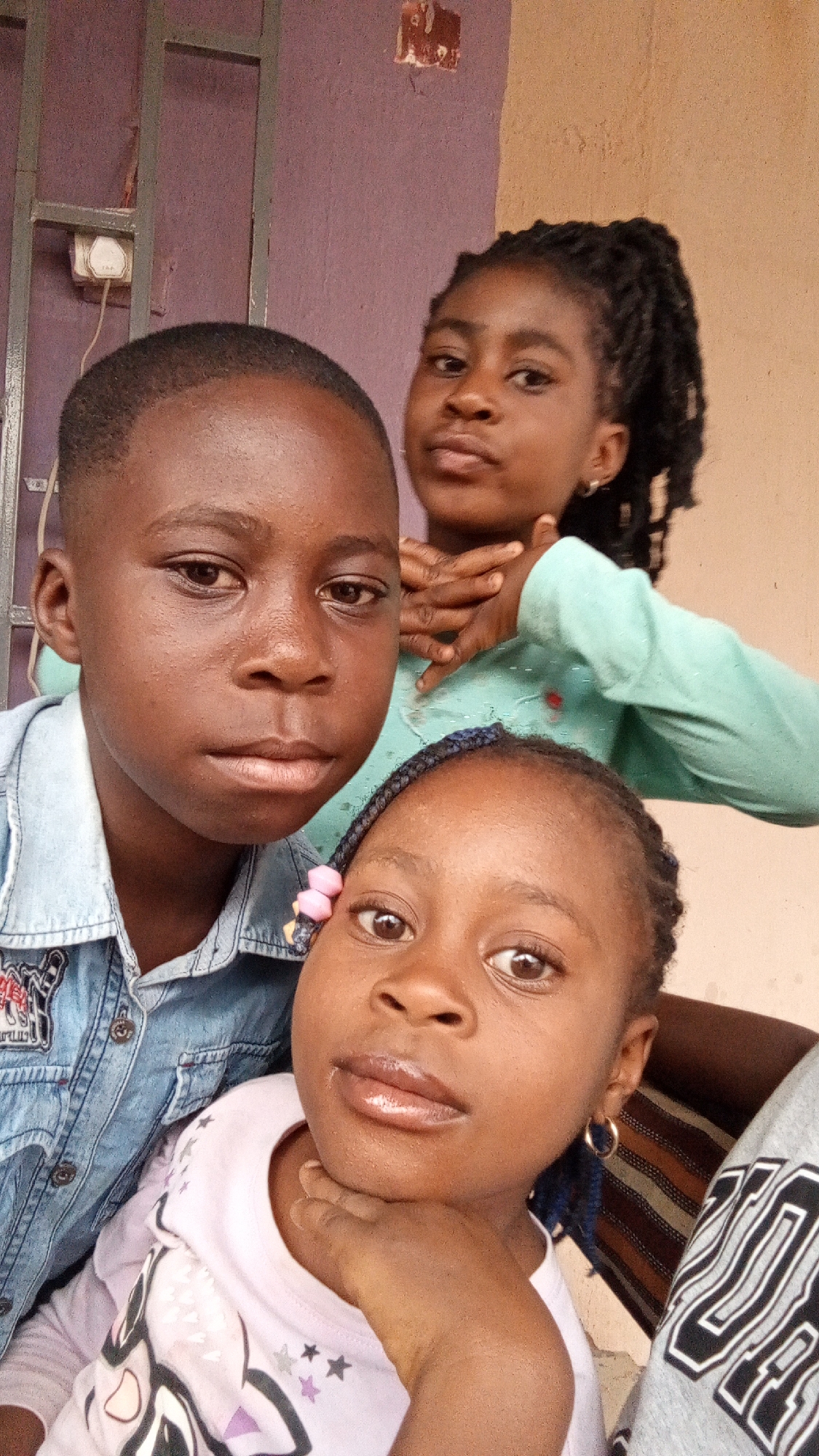 The image shows three children posing for a photograph. The location is Sabon Tasha, Nigeria.
In the foreground, a boy with short hair is wearing a denim jacket. Below him, a girl with braids and a pink hair accessory is resting her chin on her hand. Behind them, another girl with dreadlocks is holding her hands crossed in front of her chest. The background includes a section of a purple wall with an electrical outlet and a peach-colored wall.