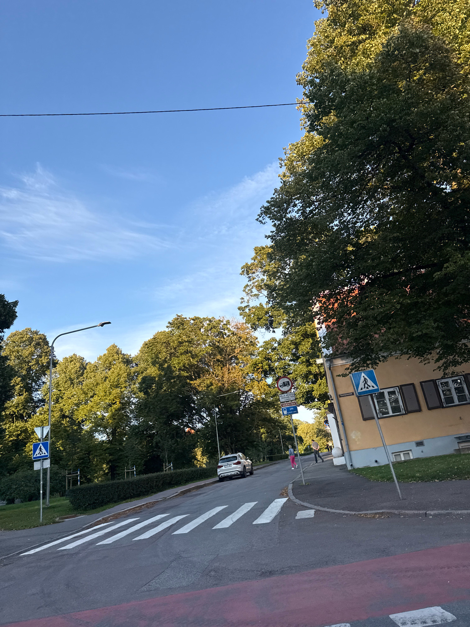 This image depicts a street scene in Tallinn, Estonia, likely on a clear day with a bright blue sky and scattered white clouds. A prominent feature is a pedestrian crossing with white stripes marked on the asphalt road. To the right of the crossing, a yellow building with brown shutters and multiple windows stands adjacent to a sidewalk. Two pedestrian crossing signs are visible; one is a blue square with a white pedestrian silhouette, and another is a circular red and white sign with text and symbols. A street lamp with a curved arm extends over the road. Trees with lush green foliage line the street and surround the buildings, casting shadows. A white car is parked on the right side of the road, and two individuals are walking on the sidewalk. The road appears to be on a slight incline.