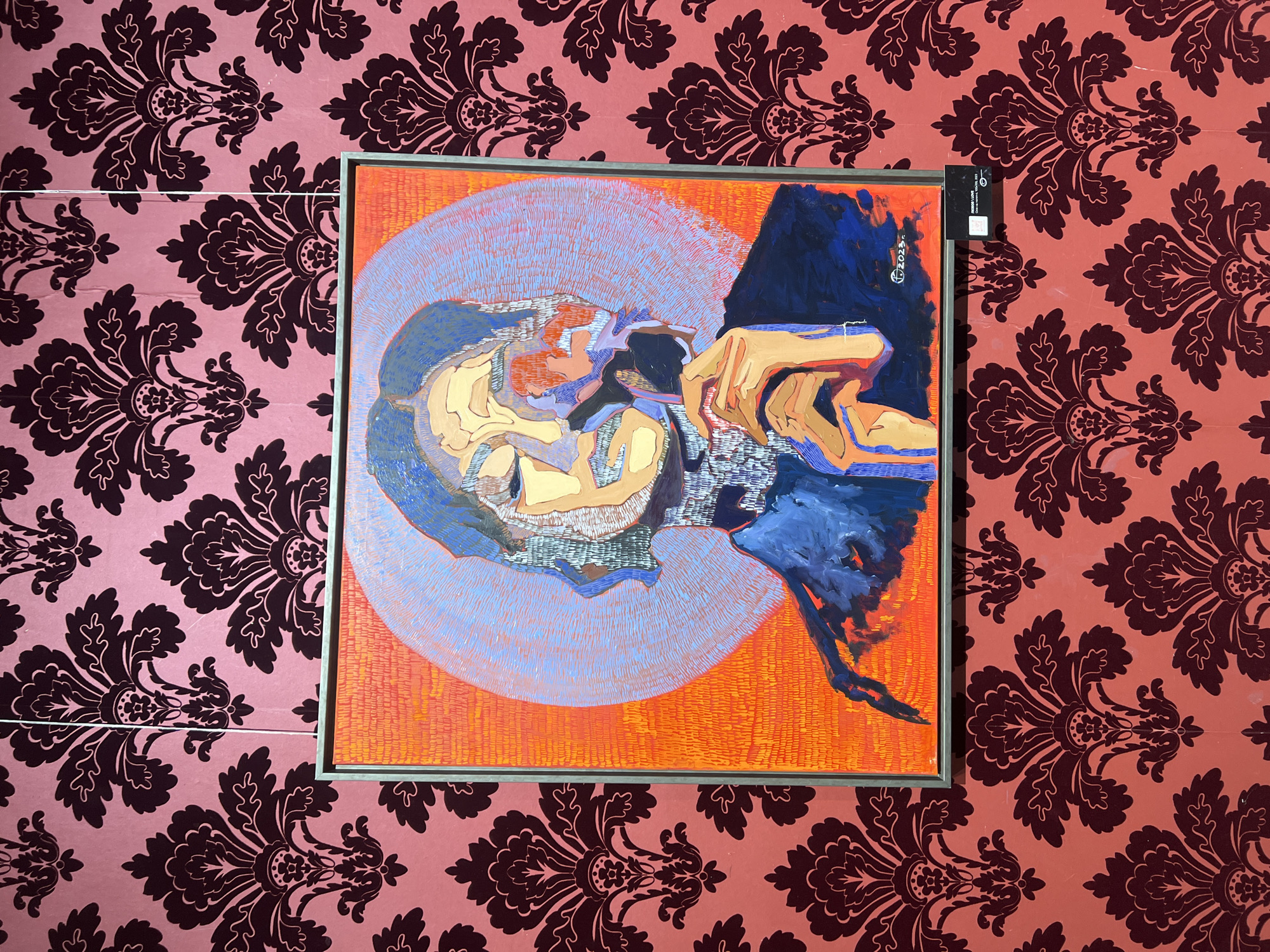 This is a medium shot photograph of a painting hanging on a wall. The painting is framed and has a silver-colored frame. The artwork itself is abstract and appears to depict a human figure or figures. There are bold brushstrokes and a vibrant color palette, with prominent use of orange, blue, and yellow. The background of the painting is a textured orange, with a circular gradient of blue in the upper left quadrant. A dark blue shape dominates the right side of the painting, with a human-like hand rendered in shades of orange and yellow reaching from it.
The wall behind the painting is covered in a repeating damask wallpaper pattern. The wallpaper is a pinkish-red color with a darker maroon or deep purple pattern.
On the right side of the painting's frame, a black label is attached. Visible text on this label includes "OOODAL LINE" and possibly "CRIAE LUBE." There are also some smudged numbers and symbols, and "2023" is visible in white on the lower left corner of the painting itself, suggesting it was created in that year. The scene is indoors, likely in a gallery or home setting. No specific time of day or weather conditions can be inferred. The emotional tone of the painting is difficult to determine definitively due to its abstract nature, but the colors and composition could suggest intensity or drama. The location context is Odesa, Ukraine, but no specific visual cues confirm this within the image itself.