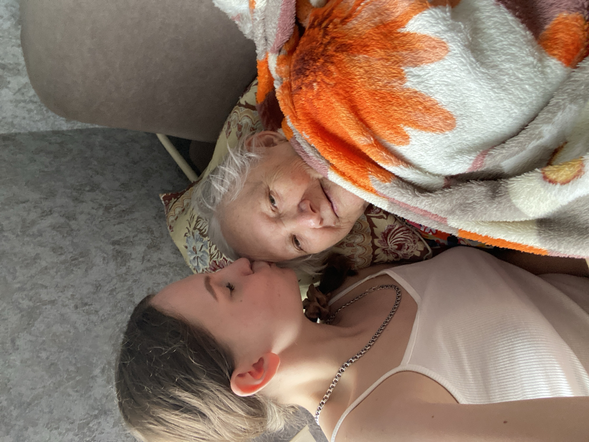 The image captures two females of different generations in an indoor setting in Марковский, Russia. On the left, a younger female, appearing to be in her late teens or early twenties, is depicted with light brown hair and wearing a white ribbed spaghetti-strap tank top and a silver chain necklace. She is engaged in the act of kissing the forehead of an older female, with her eyes closed. The older female, visibly elderly with white-grey hair, is positioned on the right, reclining against a patterned pillow featuring floral motifs. She is partially covered by a multi-colored, textured blanket displaying patterns in orange, white, and beige. The older female is looking towards the viewer with a gentle smile. The background includes a textured grey wall on the upper left and a section of light-colored upholstered furniture on the upper right.