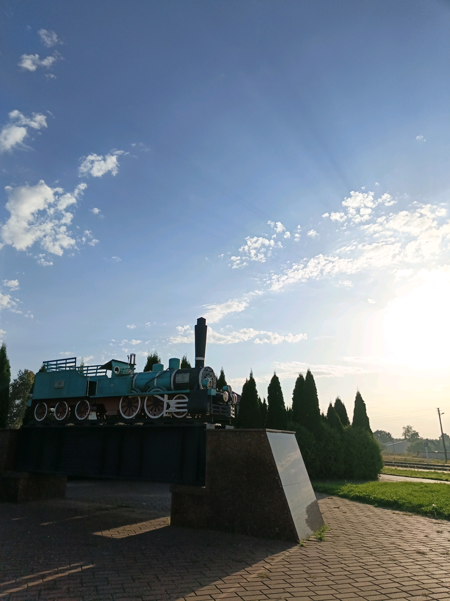 A teal and black steam locomotive monument is displayed on a dark grey pedestal in an outdoor park setting. The locomotive is positioned in the center of the frame, facing towards the right. It features white-rimmed wheels and a prominent black smokestack. Behind the locomotive, a line of dark green evergreen trees forms a backdrop. To the right of the trees, a hint of a railway track and distant buildings can be seen. The ground in the foreground is paved with reddish-brown bricks, with shadows cast by the monument and trees. The sky above is a vibrant blue with scattered white clouds, and the sun is visible in the upper right corner, casting light rays through the clouds. The scene appears to be located in the city of Porechye, Belarus.
