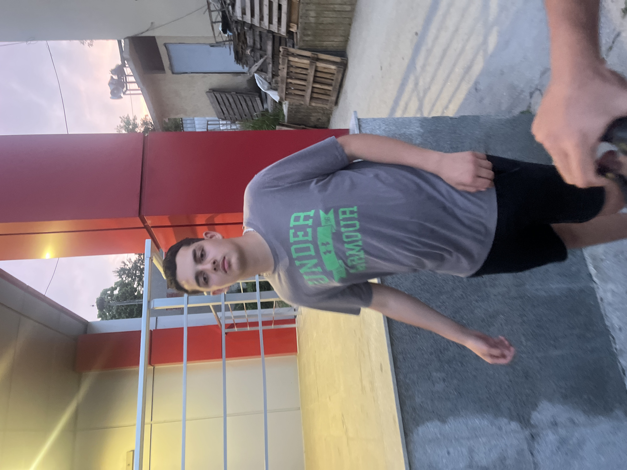 A young male, possibly in his late teens or early twenties, stands centered in the frame, facing forward with a neutral expression. He has short, dark hair and is wearing a grey short-sleeved t-shirt with "UNDER ARMOUR" in green text and a logo, and black shorts. A silver chain is visible around his neck. He stands on a paved surface, with a light-colored, possibly tiled, floor area behind him, enclosed by white and grey metal railings. To his left is a structure with white and red paneling, and a bright light source is visible on the ceiling of an overhang. To his right, an older, light-colored building with a blue door is visible, along with stacks of wooden pallets and other debris. Above the older building, on its roof, are two large, grey, possibly industrial, fan-like structures. The sky is visible in the upper right quadrant, displaying a light orange and purple hue characteristic of dusk or dawn. Trees are visible in the distance. A hand holding an unidentified dark object partially obstructs the lower right corner of the image. The scene is located in Basarbovo, Bulgaria.