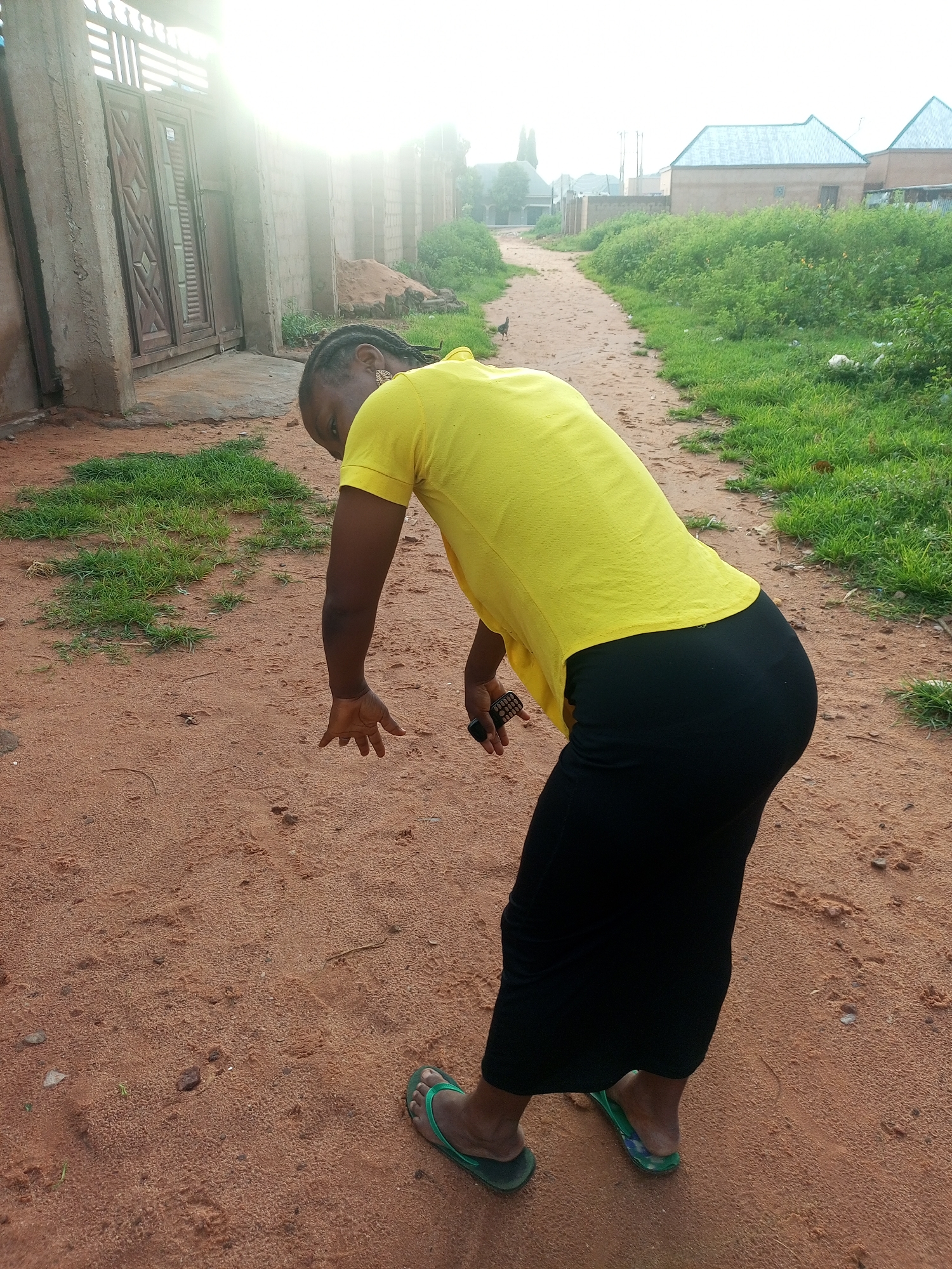 A woman wearing a yellow t-shirt and a black skirt is standing on a dirt road in Tempure, Nigeria. The woman is bent over, with her left hand reaching towards the ground. She is wearing green flip-flops. The road is lined with green grass and bushes. In the background, there are buildings with blue roofs and a fence. The sun is shining brightly, creating a lens flare in the upper left corner of the image.