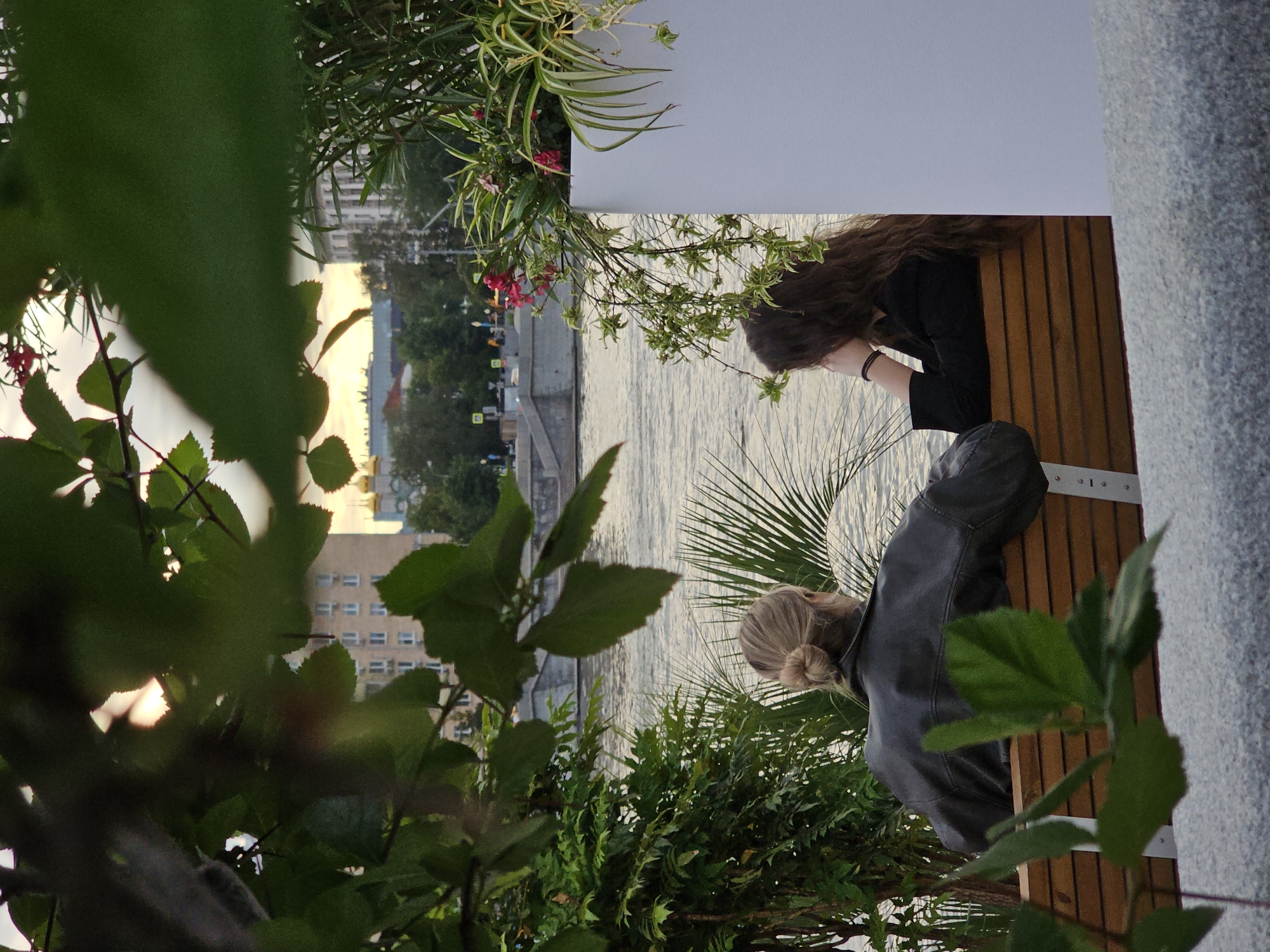 The image depicts two individuals situated near a body of water, likely a river, with a cityscape in the background. The scene is oriented sideways, with the sky appearing on the left and the ground on the right.
In the foreground, lush green foliage partially obscures the view. The two subjects are positioned on a wooden bench. The person closer to the viewer has their back turned, displaying a blonde bun hairstyle and wearing a dark leather jacket. The second individual, further away, has long brown hair and is wearing a black top. They are both looking downwards, seemingly engaged in a quiet moment or conversation.
The background reveals a river with rippling water, reflecting the soft light of what appears to be sunset or early evening. Beyond the river, buildings of various heights and architectural styles can be seen, with some lights beginning to illuminate the structures. Hints of vegetation and streetlights are also visible along the riverbank. The overall atmosphere suggests a serene, urban setting at dusk. The presence of buildings and the river suggests an urban environment, consistent with Moscow, Russia. No visible text is present in the image.
