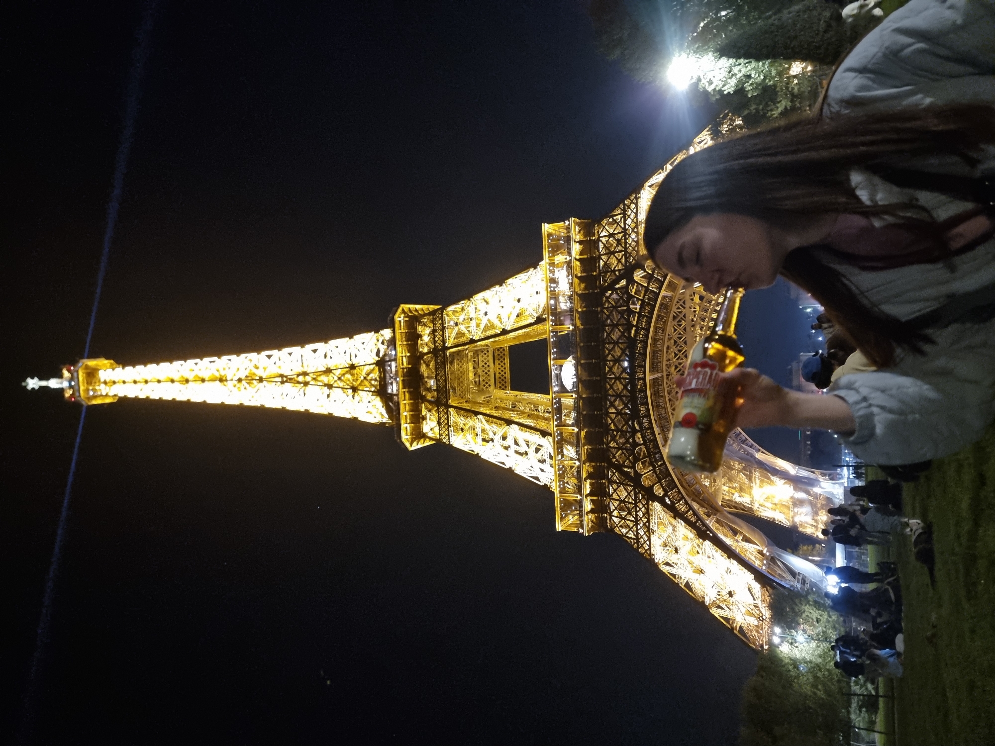 The image captures a nighttime scene in Paris, France, featuring the Eiffel Tower illuminated against the dark sky. A woman is positioned in the foreground, holding a bottle near her mouth as if drinking. Several people are gathered on the grass behind her.