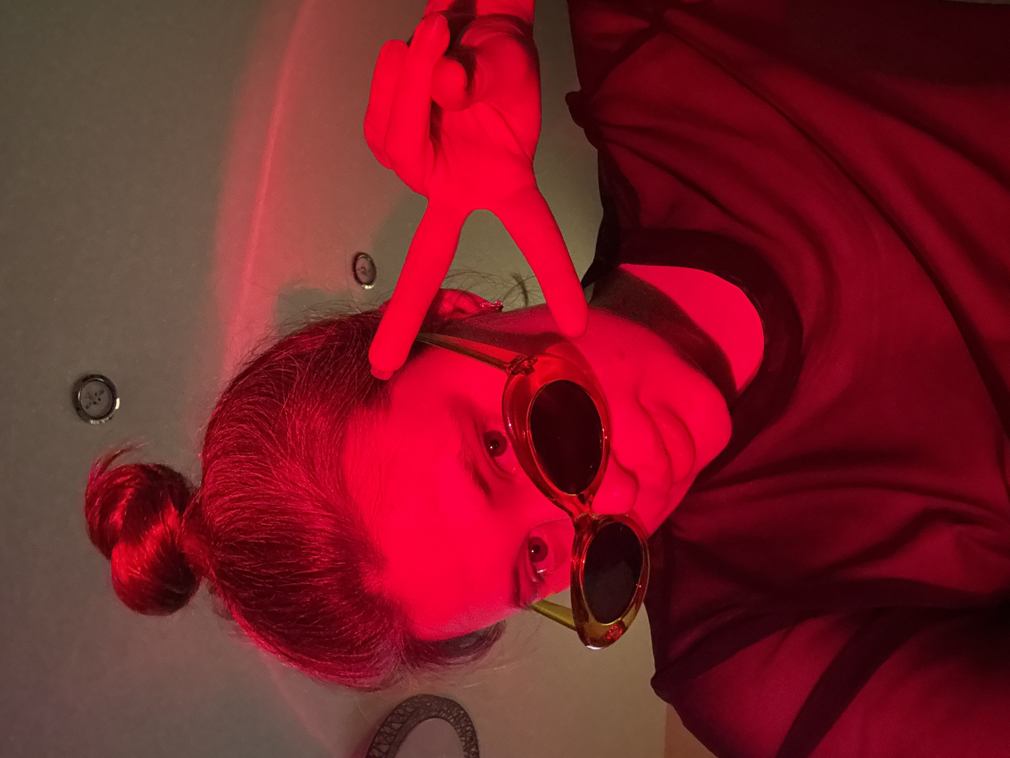 A person with their hair in a bun is photographed from below while wearing sunglasses and making a peace sign with their hand. The scene is illuminated with a red light. The setting is a light-colored room, likely a bathroom due to the presence of what appears to be a jacuzzi tub. The location cannot be determined, especially not the city of Chaikovsky, Russia.