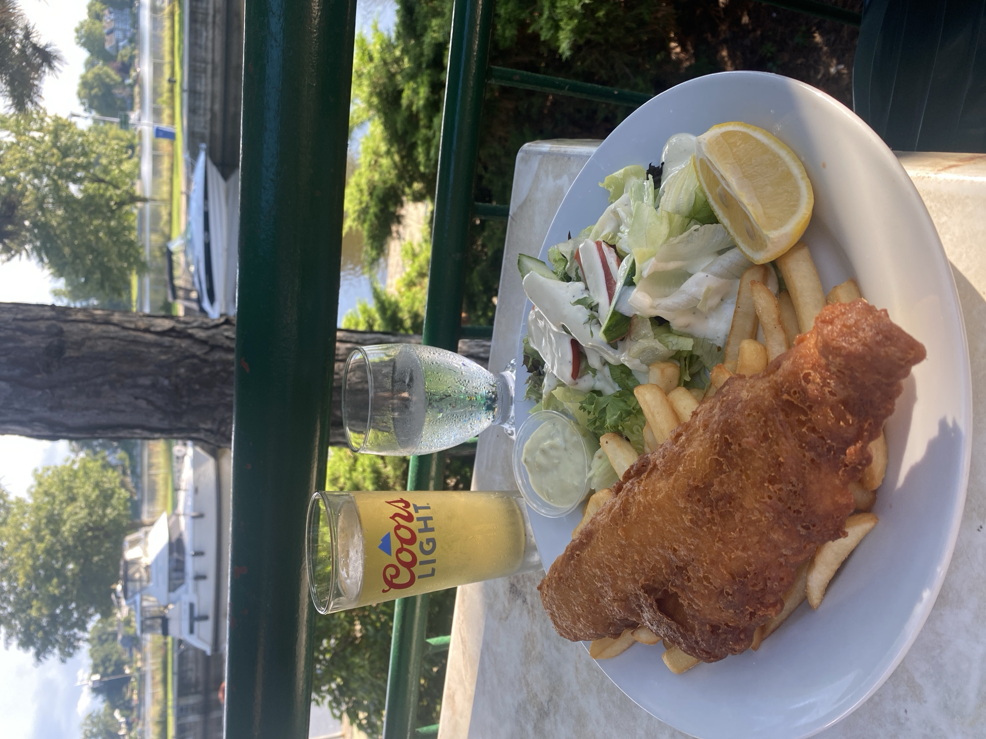 The image depicts a plate of fish and chips served with a side salad, tartar sauce, and a lemon wedge. The meal is accompanied by a tall glass of Coors Light beer and a glass of water. The setting appears to be an outdoor patio with a view of a body of water, possibly the Ottawa River, in Sainte-Anne-de-Bellevue, Canada. Several boats are docked in the background, and lush green trees and foliage surround the area, suggesting a sunny day with good weather. The table is covered with a light-colored, speckled surface. The railing in the foreground is painted dark green.