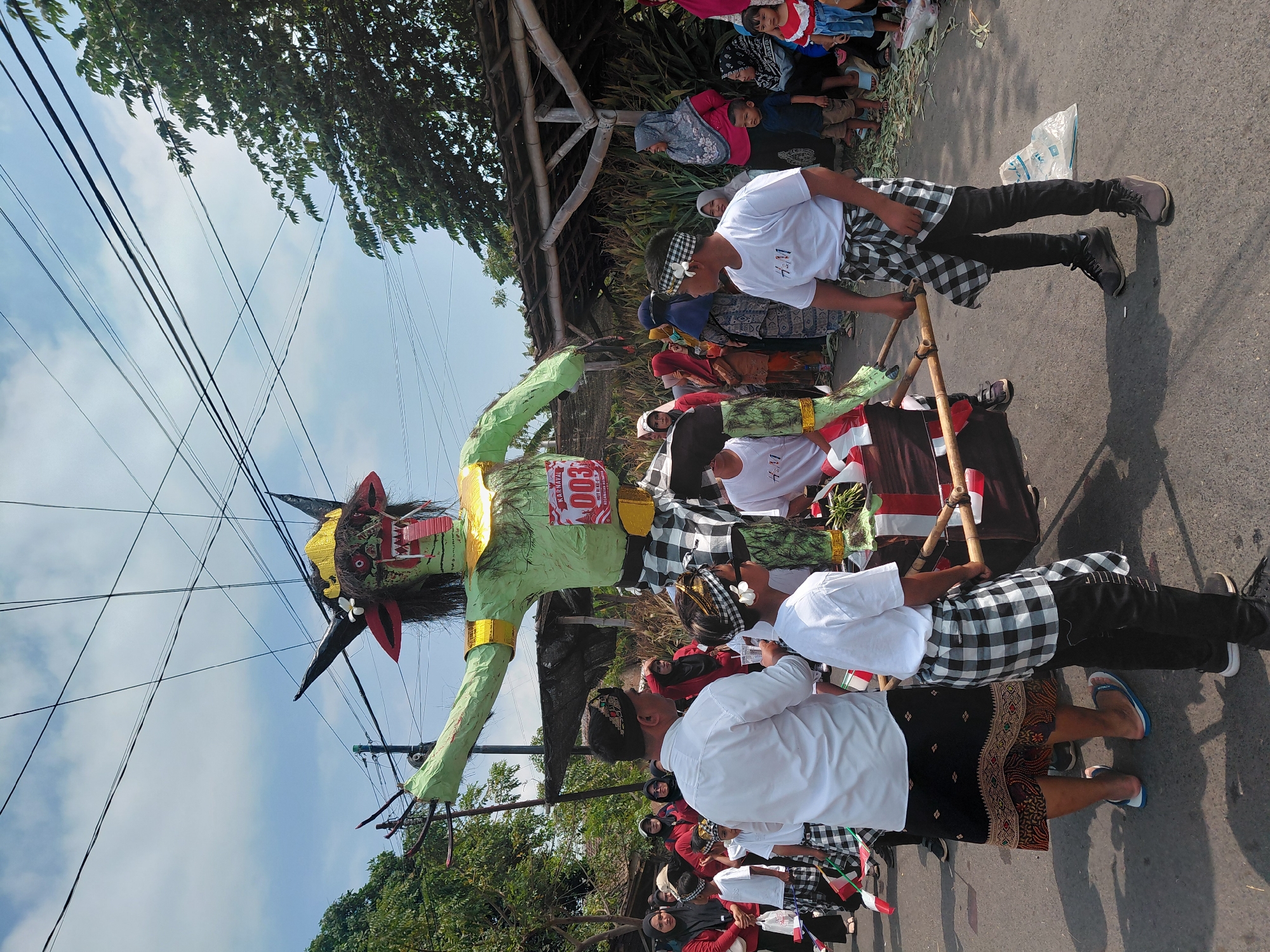 In the city of Bimo, Indonesia, a procession is underway, likely a carnival or festival, given the presence of a large, green, monstrous effigy being carried by several individuals. The effigy, which resembles a mythical creature with horns, sharp teeth, and a protruding red tongue, is adorned with golden accents and a sash bearing the number "003" and the text "KARNAVAL." A crowd of onlookers, dressed in a mix of traditional and modern attire, observes the event. Many of the participants, especially the younger ones, are wearing checkered black and white sarongs, a common cultural garment. The scene takes place on a street, with trees and some makeshift shelters visible in the background. The sky is partly cloudy, suggesting a warm day. The overall atmosphere appears festive and communal, with a focus on the ceremonial transport of the effigy.