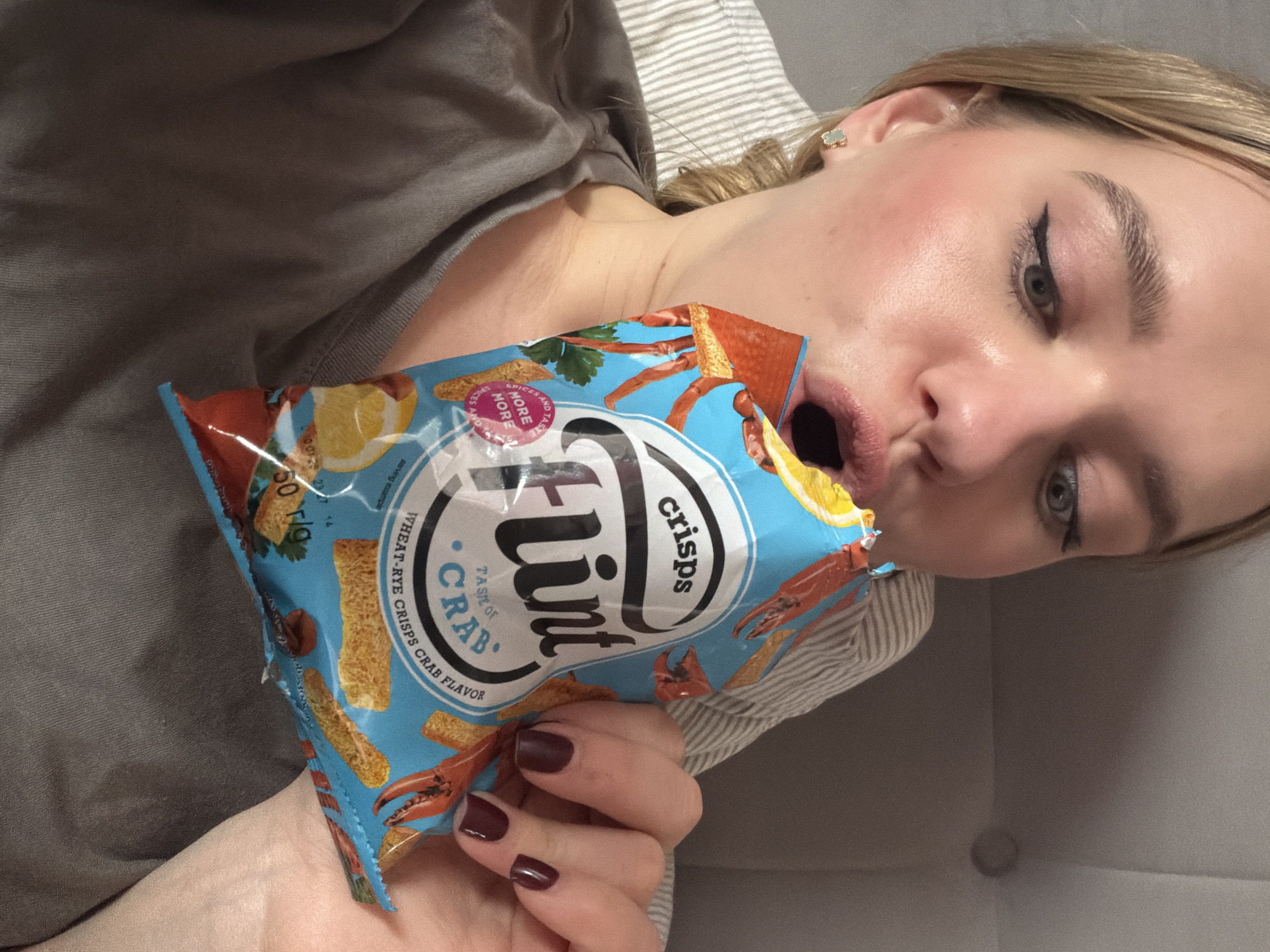 A fair-skinned woman is lying down, possibly in bed, angled towards the camera, with her face close to the lens. She has light blonde hair, subtle makeup including winged eyeliner, and is wearing a grey top. She holds a blue packet of "Flint crisps" with "Taste of Crab" flavor up to her mouth, which is open in what appears to be a playful or exaggerated expression. Her nails are painted a dark reddish-brown. The crisp packet is adorned with images of lemons and crab claws. The packet also says "Wheat-Rye Crisps Crab Flavor". The background is blurred, but suggests a striped pillow and a grey upholstered surface. The image was taken in the city of Dreiliņi, Latvia.