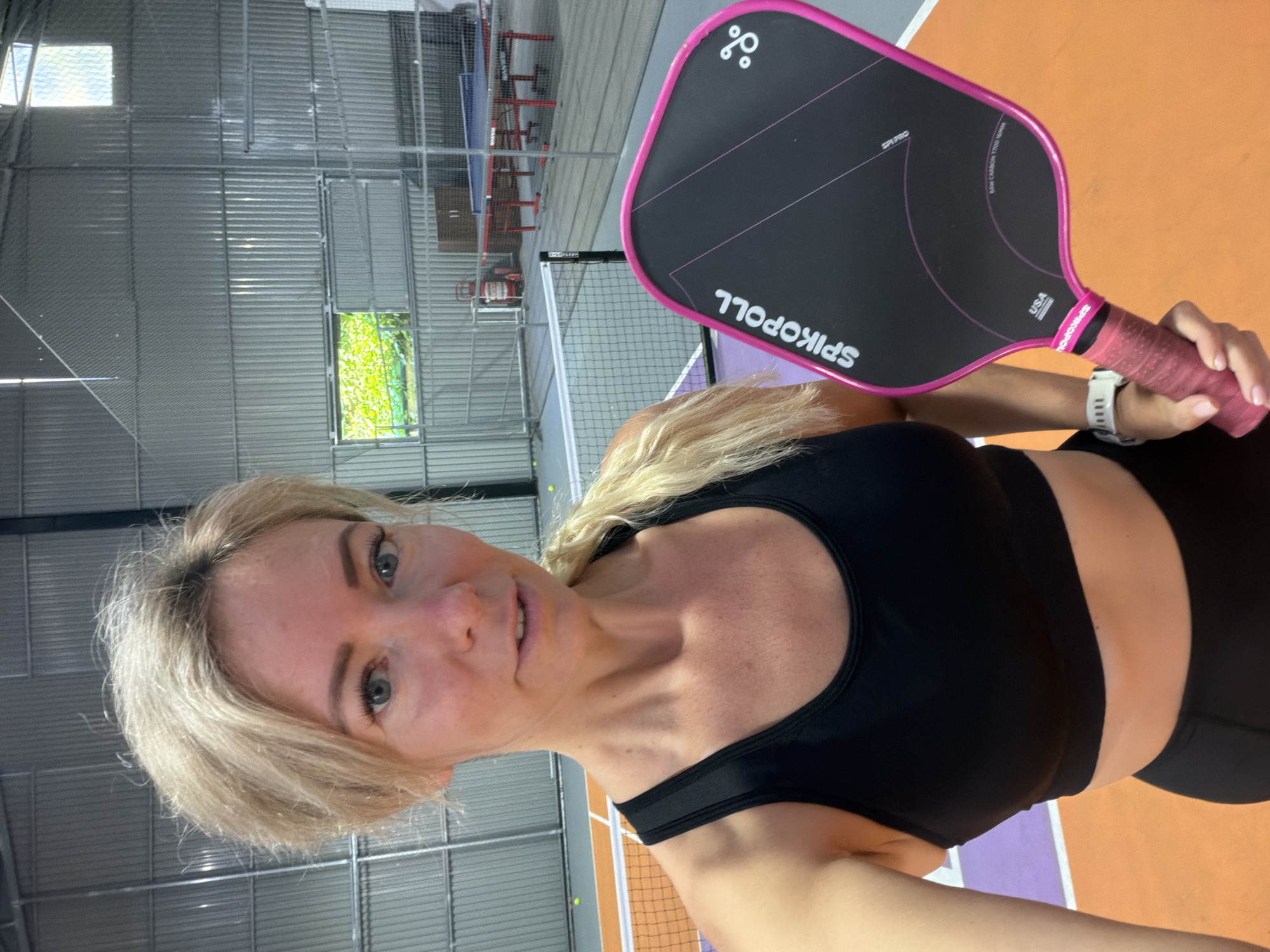 A woman with blonde hair pulled back in a ponytail, wearing a black sports bra and black leggings, holds a pink and black pickleball paddle. She is standing on an orange and purple pickleball court indoors. In the background, there are pickleball nets, ping pong tables, and windows showing greenery outside. The walls of the indoor facility are made of corrugated metal.