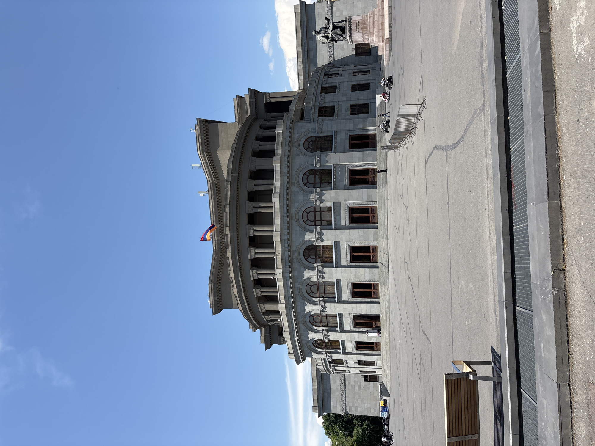 The image captures a wide shot of the Yerevan Opera House, a prominent landmark in Yerevan, Armenia, on a clear day with a bright blue sky. The grand stone building, characterized by its neoclassical architecture, features a semicircular facade with arched windows and a colonnade. An Armenian flag flies atop the building. In front of the opera house is a large, paved plaza. To the right of the building, a bronze statue of a seated figure is visible. Several people are present in the scene, including individuals walking and riding ATVs in the plaza. A wooden bench is situated in the foreground, to the left of the frame. The overall scene depicts a public space in front of an important cultural institution.