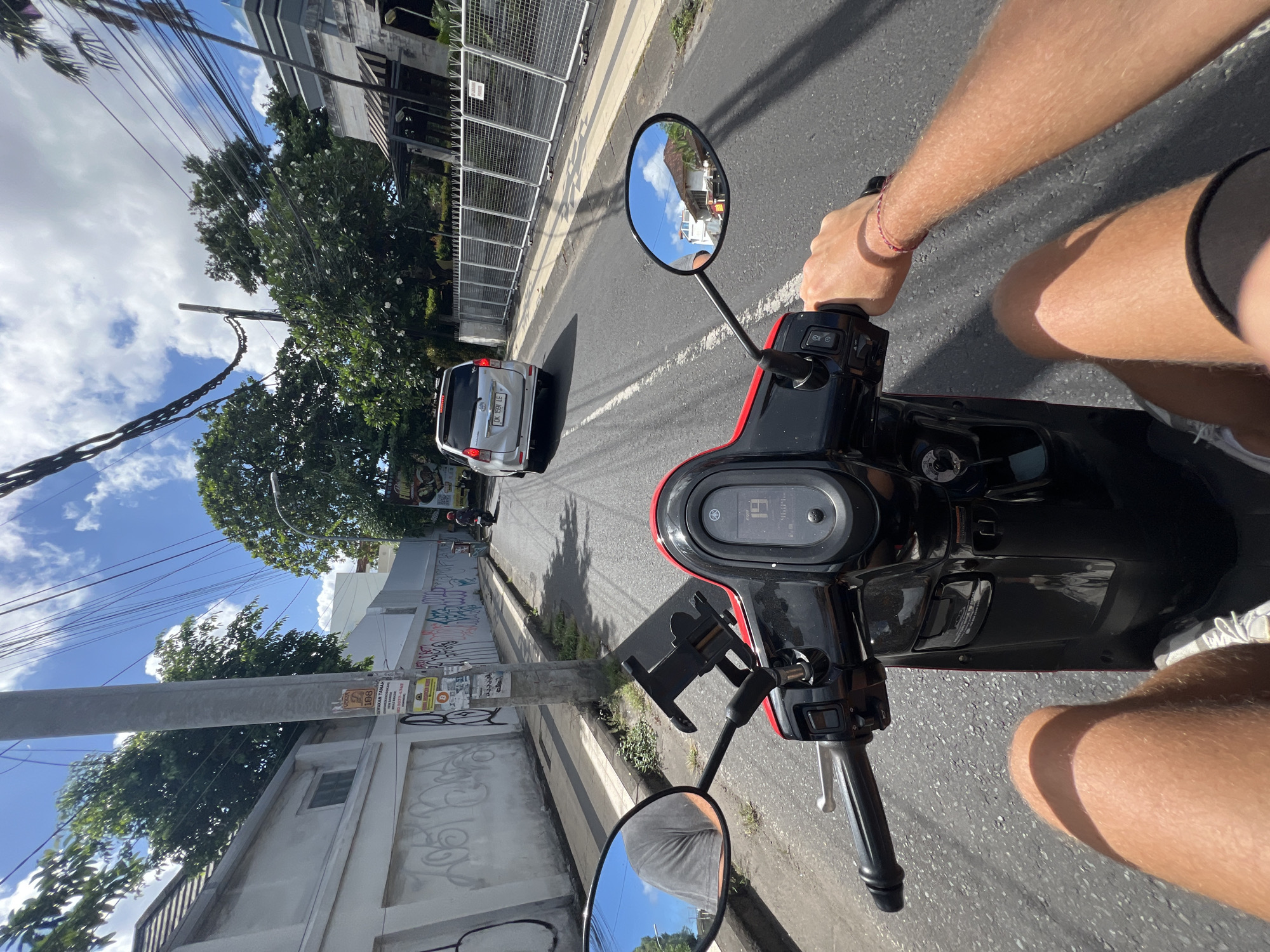 A street-level perspective from the point of view of someone riding a black and red scooter on a paved road in Kuta, Indonesia. The rider's bare arm and thigh are visible on the right side of the frame, and the scooter's handlebars, mirrors, and instrument panel are prominent in the foreground. The speedometer reads 19. A silver car is ahead on the road, and buildings, trees, and power lines line the sides of the street. Graffiti is visible on a wall to the left. The sky is partly cloudy.