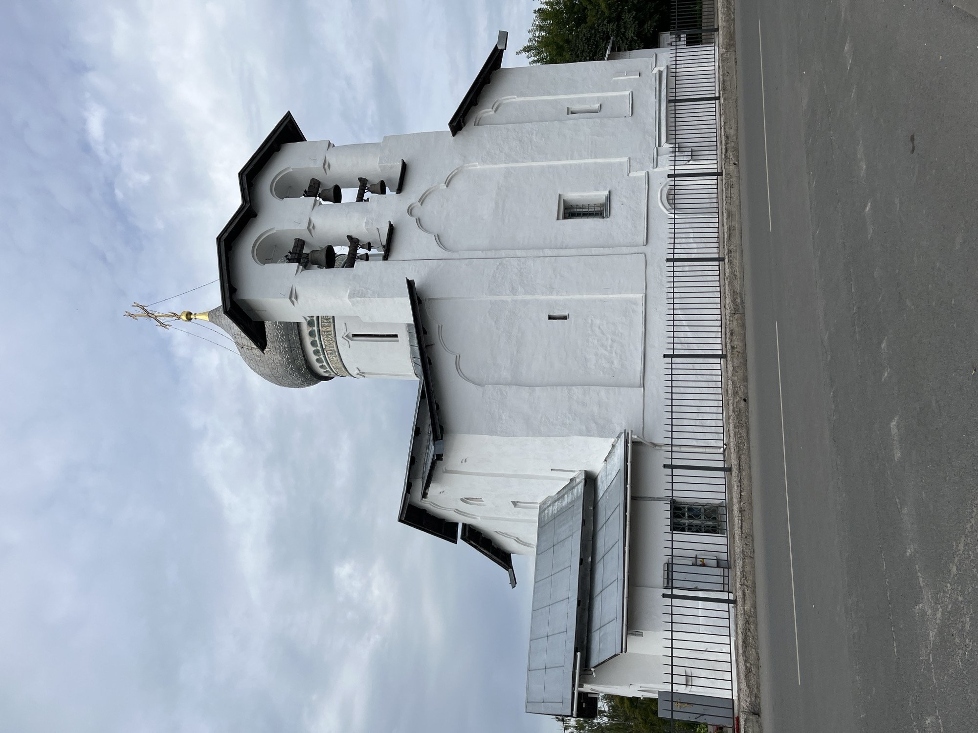This is a side-view, eye-level, medium shot of the Church of the Transfiguration of the Savior on Ilyina Street in Pskov, Russia. The church is painted white with a dark gray, multi-faceted dome topped with a golden cross. Attached to the main building is a white bell tower with arched openings, containing several bells. The church is surrounded by a black, spiked metal fence, and in front of the fence is a paved road with a white dividing line. The sky is overcast with a mixture of light and dark gray clouds. No people are visible in the image.