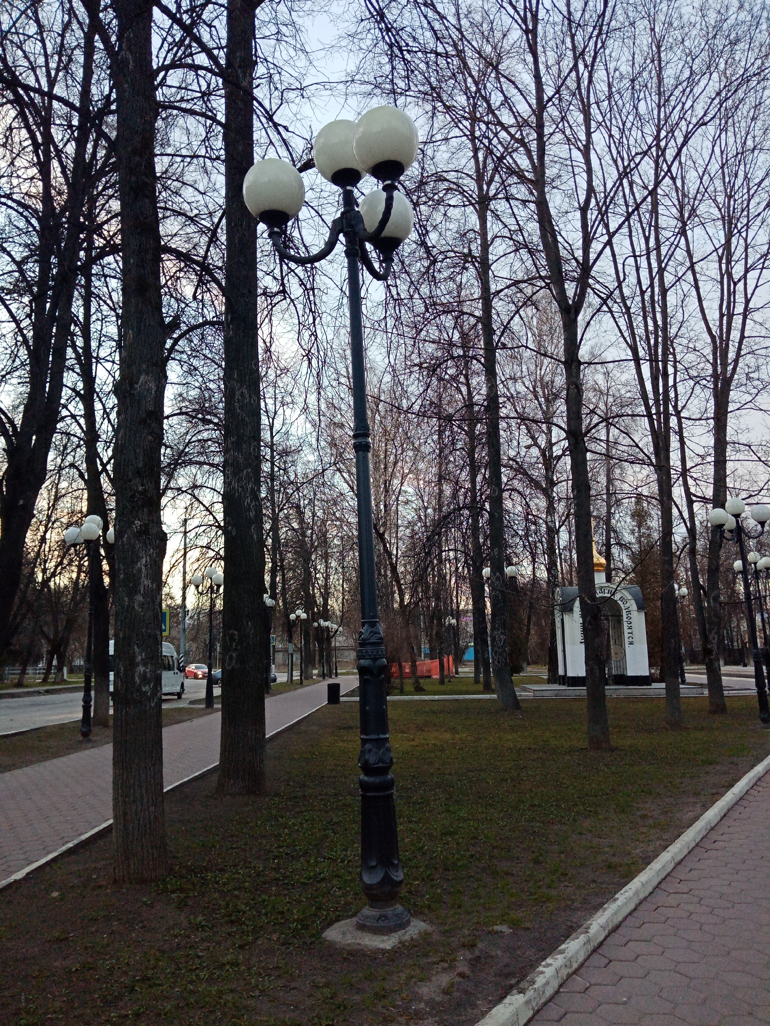 The image depicts a park scene in Elektrostal, Russia, characterized by a paved walkway bordered by bare trees and grass patches. A prominent feature is an ornate black lamppost with four globe-shaped lights. In the background, a small white chapel with a gold dome is visible among the trees. The sky appears overcast, and a few vehicles can be seen on a road in the distance. Additional lampposts line the walkway.