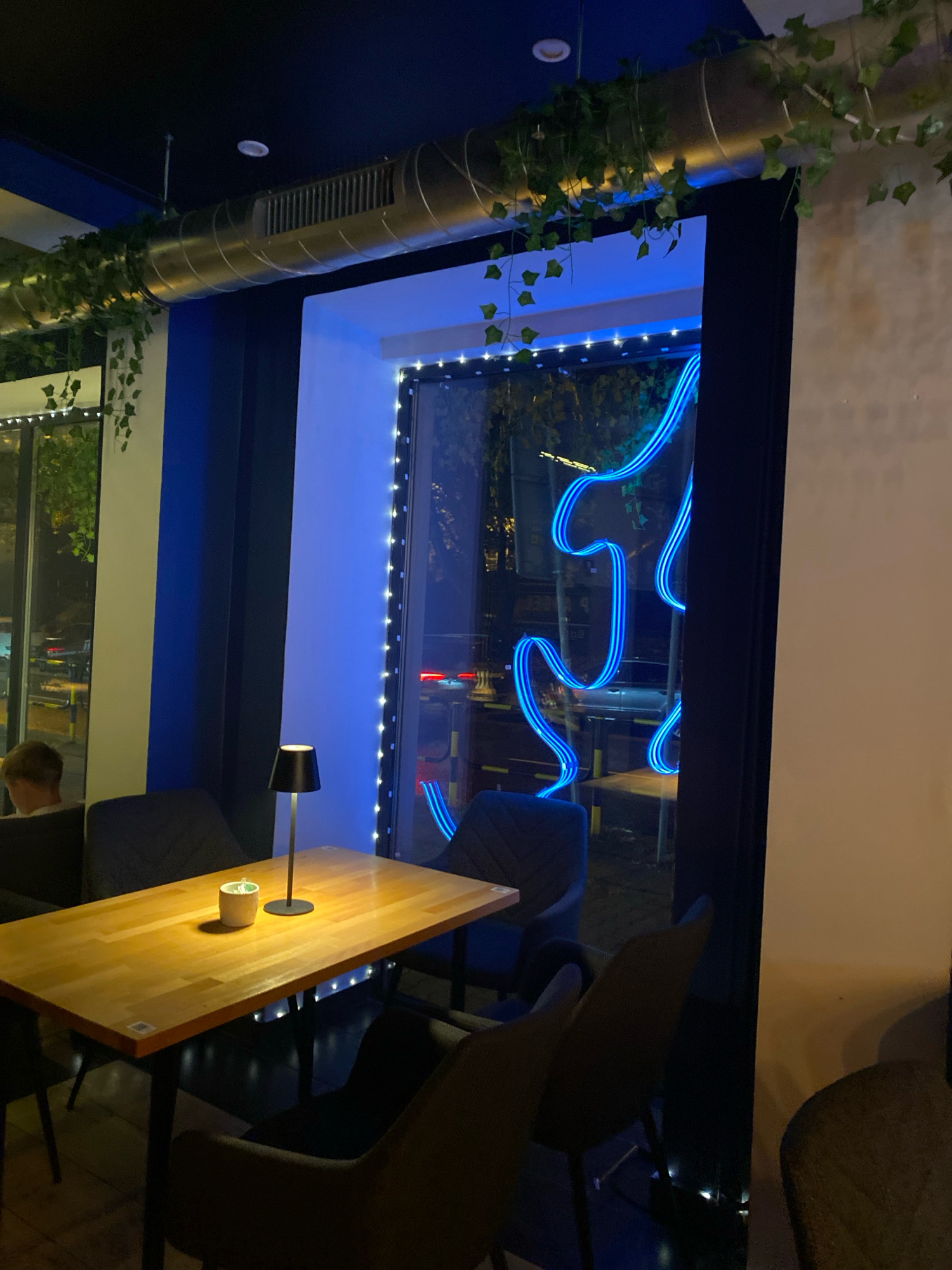 The image shows a dimly lit interior, possibly a cafe or restaurant, with a large window displaying a neon blue light sculpture. The window is framed by black curtains and adorned with a string of white fairy lights. Outside the window, streetlights and building lights are visible, suggesting it is nighttime.
Inside, a light wooden table is set with a small black lamp and a white plant pot. Several chairs with dark upholstery surround the table. A person with blond hair is partially visible sitting in a chair on the left side of the frame, facing away from the camera. Above the window, a silver ventilation duct is decorated with artificial green ivy. The walls are painted in dark blue and white.
Based on the available information, this scene is located at the "Cafe & Bistro Śląskie Smaki" at ul. Bytomska 22, 44-100 Gliwice, Poland.