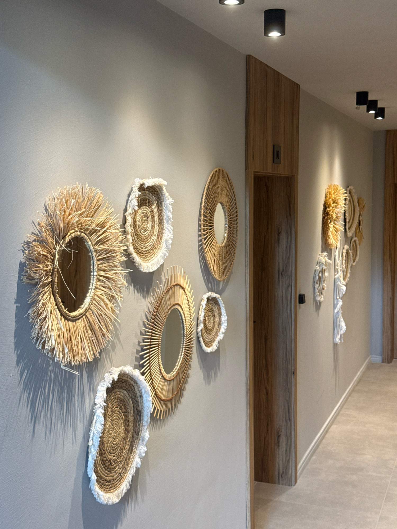 This is a full-frame, eye-level, medium-wide shot of a hallway interior in Sozopol, Bulgaria. The hallway features a light grey wall adorned with several round, decorative wall hangings made of natural materials like straw and woven fibers. Some of these hangings are mirrors with decorative frames. To the right of the wall hangings is a wooden door frame with a dark wood door. The ceiling has multiple black cylindrical lights. The floor is tiled with light beige rectangular tiles. The overall scene suggests a modern and minimalist interior design with a touch of bohemian or natural aesthetic.