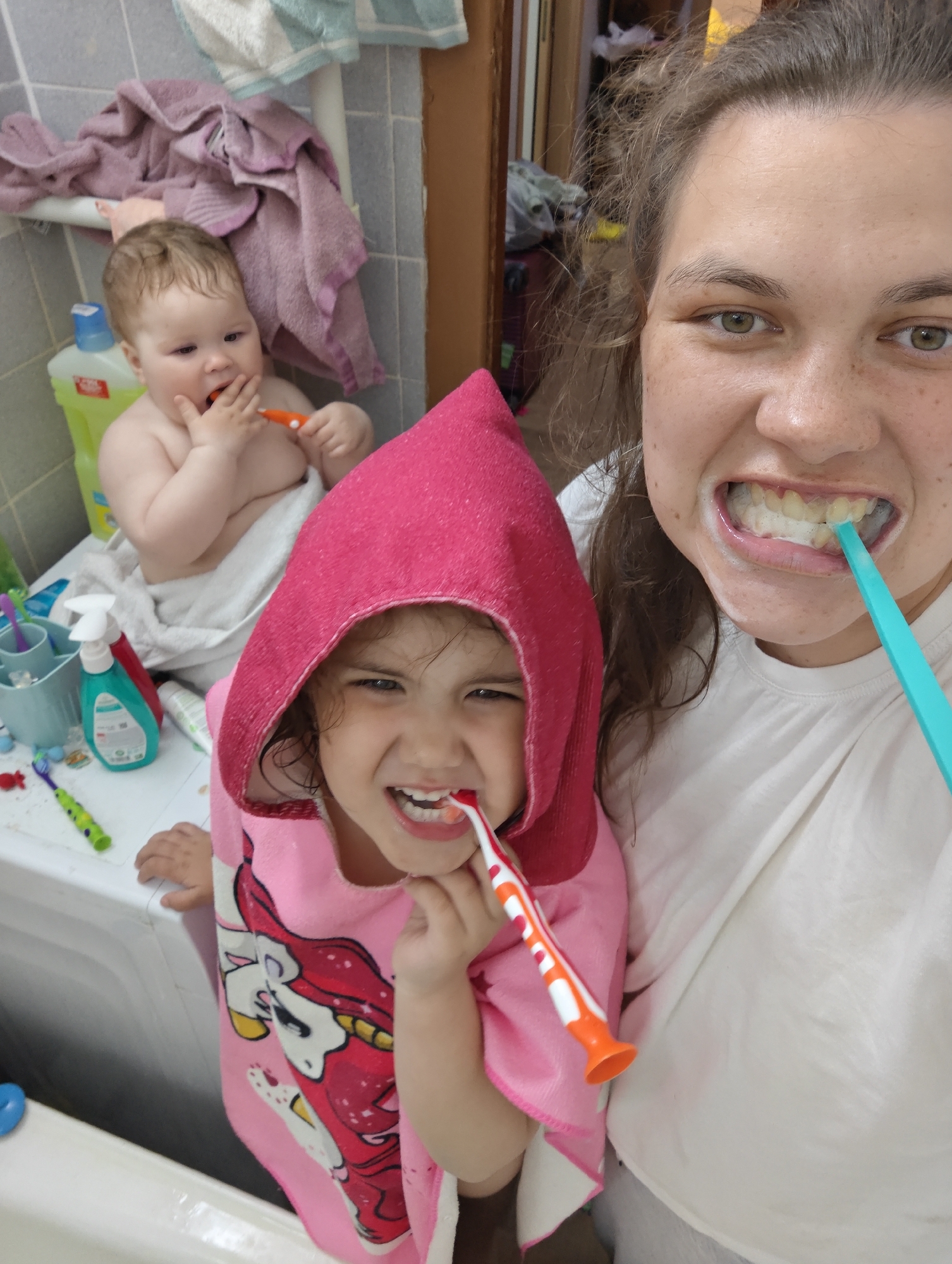 The image depicts a woman and two young children in a bathroom setting. The woman is holding the camera and smiling, with a blue toothbrush in her mouth. A younger child in a pink hooded towel stands next to her, also brushing teeth with an orange and white toothbrush. In the background, a baby sits on a white surface, draped in a towel, and also holding a toothbrush. Various bathroom items are visible on the surface where the baby sits, including toothpaste, cleaning supplies, and toothbrush holders. Towels are draped over a rack in the background. The location is potentially Chaikovsky, Russia, given the user's request, although there are no specific landmarks or signs within the image to confirm this. The scene suggests a moment of familial hygiene, with the adults and children engaged in brushing their teeth together.