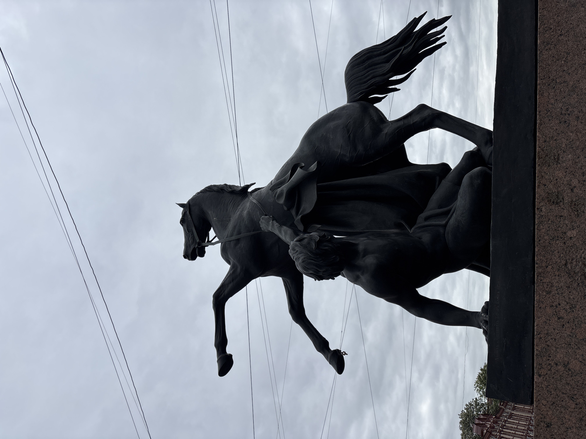 A low-angle, oblique photograph captures a dark-toned statue against an overcast sky, partially obscured by electrical wires. The statue depicts a rearing horse, its tail flowing, ridden by a figure that appears to be restraining or overpowering a fallen man. The base of the statue is visible on the right, showing dark and reddish-brown stone. The surrounding buildings and trees can be seen just beneath the edge of the statue's base. The composition is rotated slightly to the side, with the statue dominating the view and the sky providing a backdrop. It is the Horse Tamers sculpture at Anichkov Bridge in Saint Petersburg, Russia.