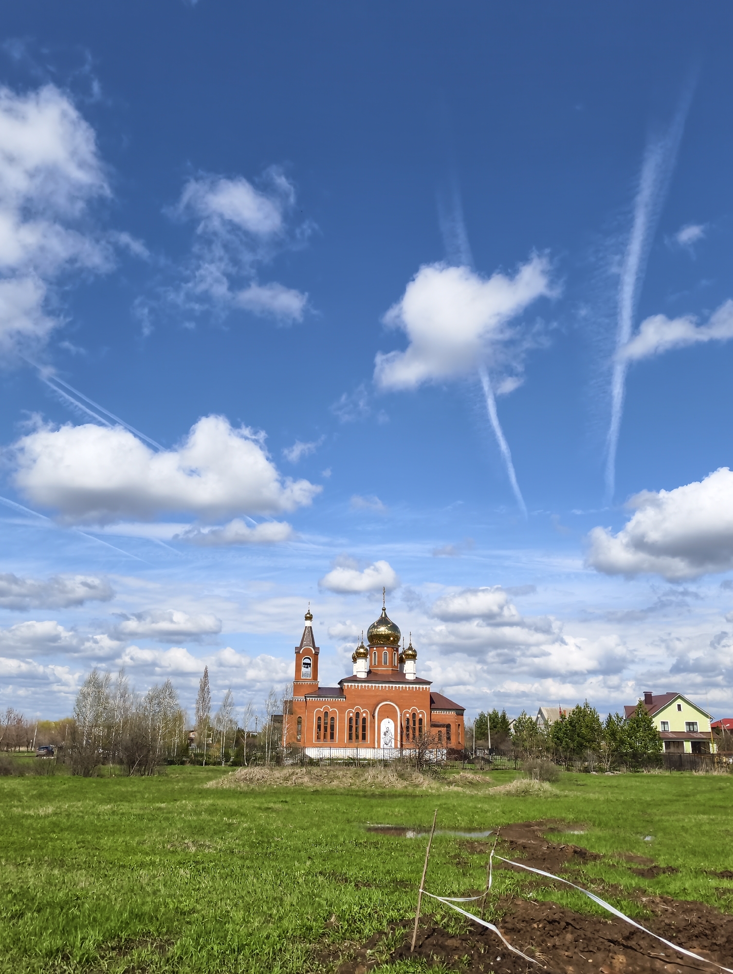 The image shows a clear, bright day with blue sky and scattered cumulus clouds marked by airplane contrails. In the center stands a red brick Russian Orthodox church with golden onion domes. The church is surrounded by a green field, with trees and houses visible in the background. In the foreground, there is disturbed soil and white tape outlining a section of the field. The setting is likely rural or suburban. Based on prior knowledge, the image is located in the city of Balashov, Russia.