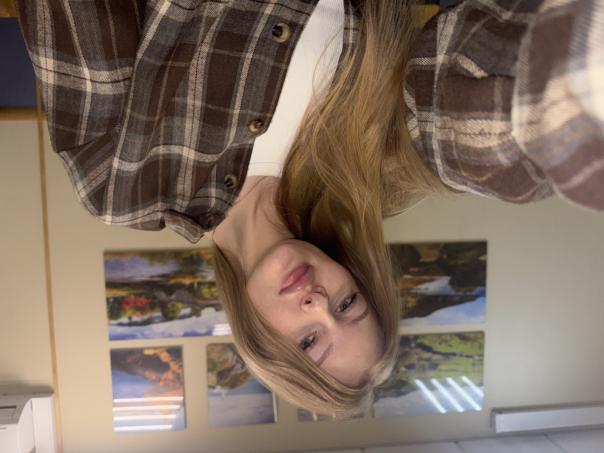 A young woman with long blonde hair is posing for a selfie indoors. She is wearing a brown and white plaid flannel shirt over a white top. In the background, there are several landscape pictures on the wall. There is also a white object that could be a printer, against the wall. The image does not contain enough information to determine the location in Perm, Russia.