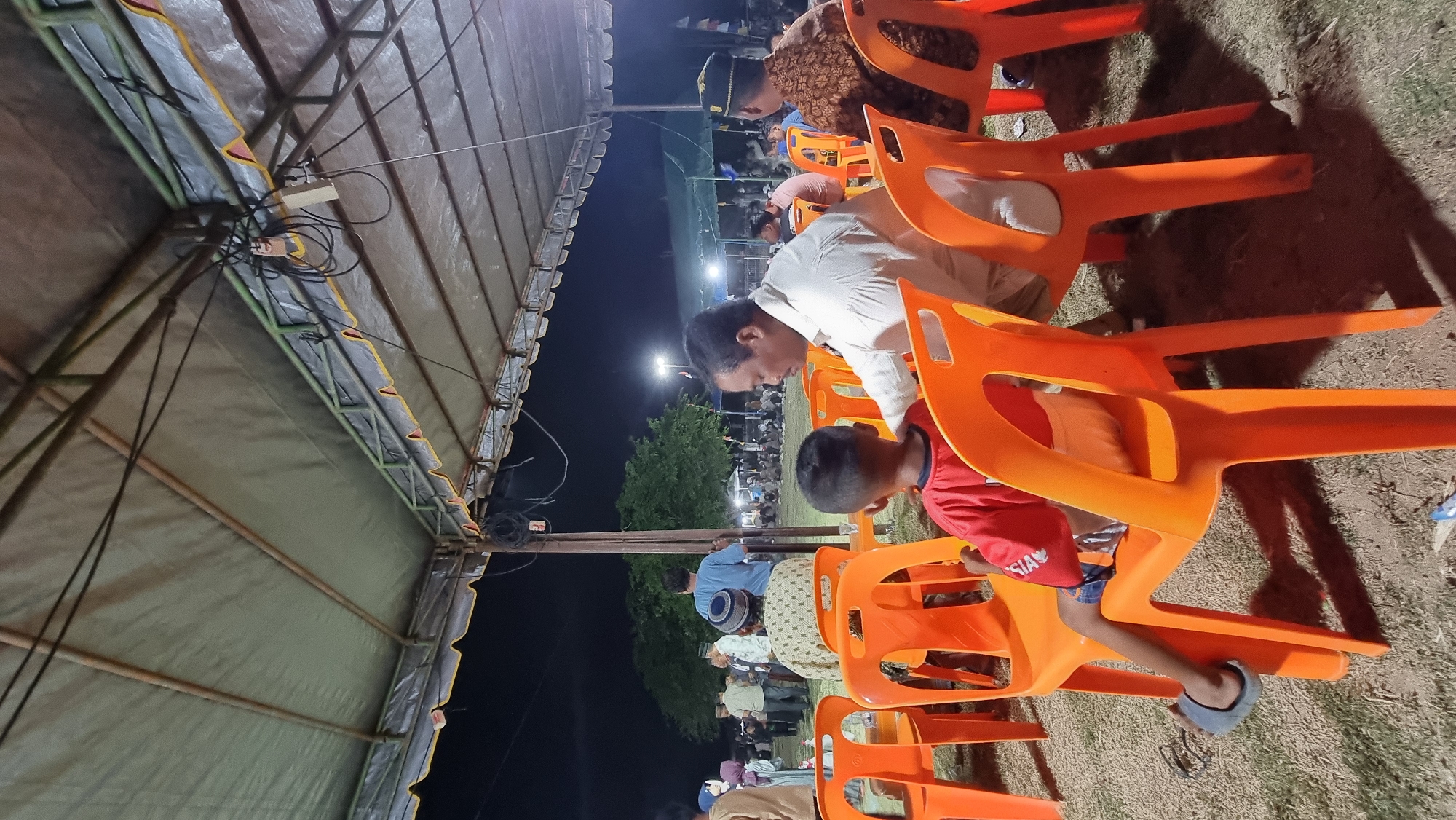 In Tumpokbeurandang, Indonesia, an outdoor event is underway beneath a large, canvas-covered tent. The tent, supported by metal poles and framing, is illuminated by overhead lights, casting shadows on the dry, grassy ground below. Several people are seated on bright orange plastic chairs, facing away from the camera. In the foreground, a young boy in a red shirt and blue shorts sits with his back to the viewer, his feet resting on the seat of his chair. To his right, a man in a white shirt is seated, looking towards something outside the frame. Further back, more individuals are visible, some seated, others standing. A cluster of trees is seen in the middle ground against a dark night sky. In the distance, additional tents and lights suggest a larger gathering or festival. The overall scene depicts a communal outdoor event in a semi-rural setting at night.