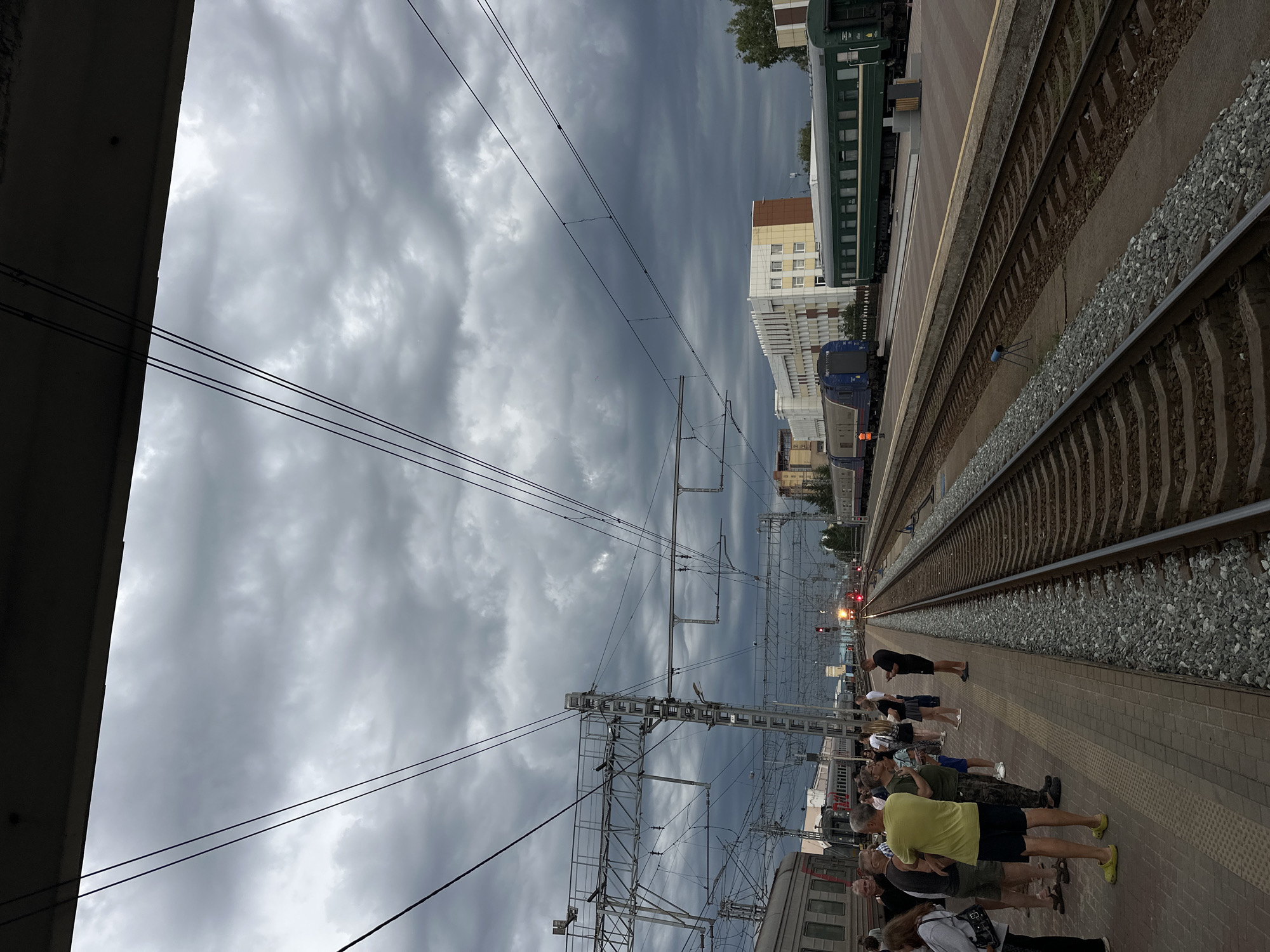The image depicts a train station platform in Omsk, Russia, under a heavily overcast and dark sky, suggesting stormy weather or late evening. Several people are standing on the platform, facing towards the railway tracks and seemingly waiting for a train or observing its approach. Some individuals are dressed in casual summer attire, including t-shirts and shorts.
On the tracks, a train is visible in the distance, with its headlights illuminated, indicating movement or readiness to depart. Another train, predominantly blue and grey, is parked closer to the foreground on the left side. The platform itself is paved with brick-like tiles, and a yellow tactile paving line runs along the edge. Overhead, complex electrical wires and support structures for an overhead railway line dominate the sky. In the background, modern buildings, including one with a beige and white facade, are visible. The overall atmosphere appears somewhat subdued, possibly due to the dark weather. No visible text is present in the image. The scene captures a moment of anticipation at a Russian train station.